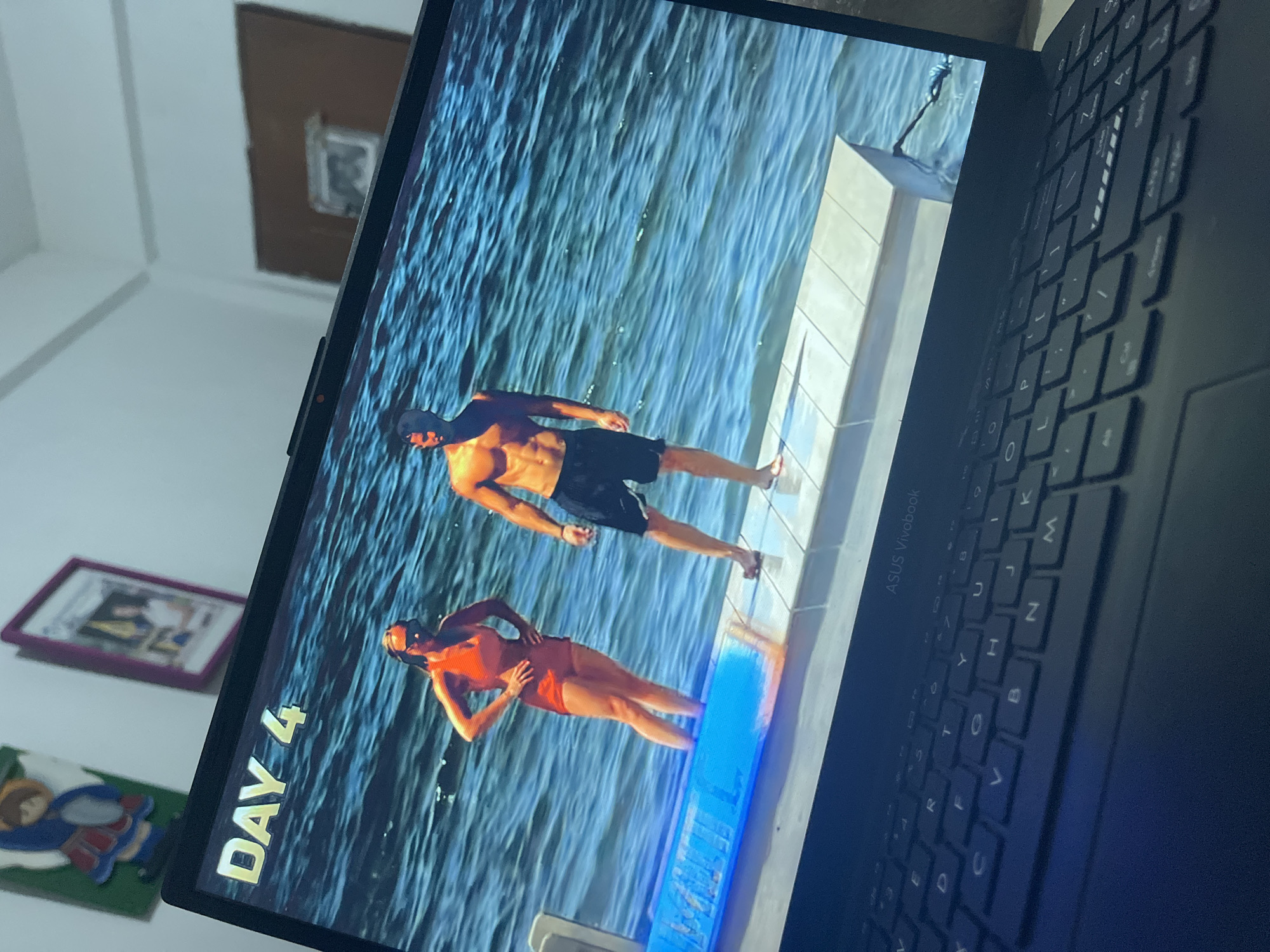 A black ASUS Vivobook laptop, partially visible from an oblique angle, displays a video featuring two individuals on a pier or platform in a body of water. The screen shows the text "DAY 4" in gold letters in the upper left corner. A female individual stands to the left, partially submerged in shallow blue water up to her shins, wearing an orange swimsuit and sunglasses, with her hands on her hips. A male individual stands to the right on the white platform, shirtless and wearing black swim shorts, looking towards the right side of the frame. The water exhibits visible ripples and waves.
The laptop's keyboard is visible in the foreground. In the background, a light-colored wall features a green-backed cutout of a cartoon-style figure wearing a blue top and red skirt on the upper left. To the upper right, a framed item with a purple border is affixed to the wall. Further right, a large brown rectangular object, possibly a door or panel, is visible against the wall. The scene is located in the city of Santa Ana, Venezuela.