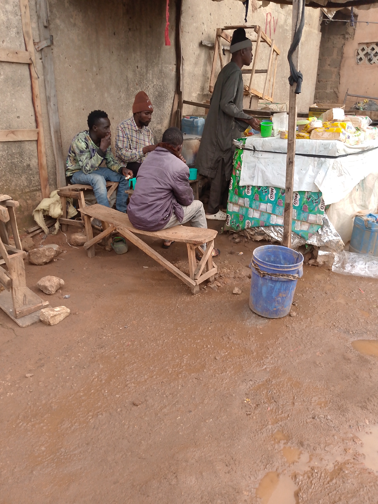 The image depicts a street scene in Bukuru, Nigeria. Three men are sitting on a rough, wooden bench next to a building with a weathered wall. A fourth man, wearing a tall hat and robe, stands at a roadside stand selling items covered with a white sheet and Milo-branded packaging. A blue bucket sits on the ground. The ground is dirt with puddles.