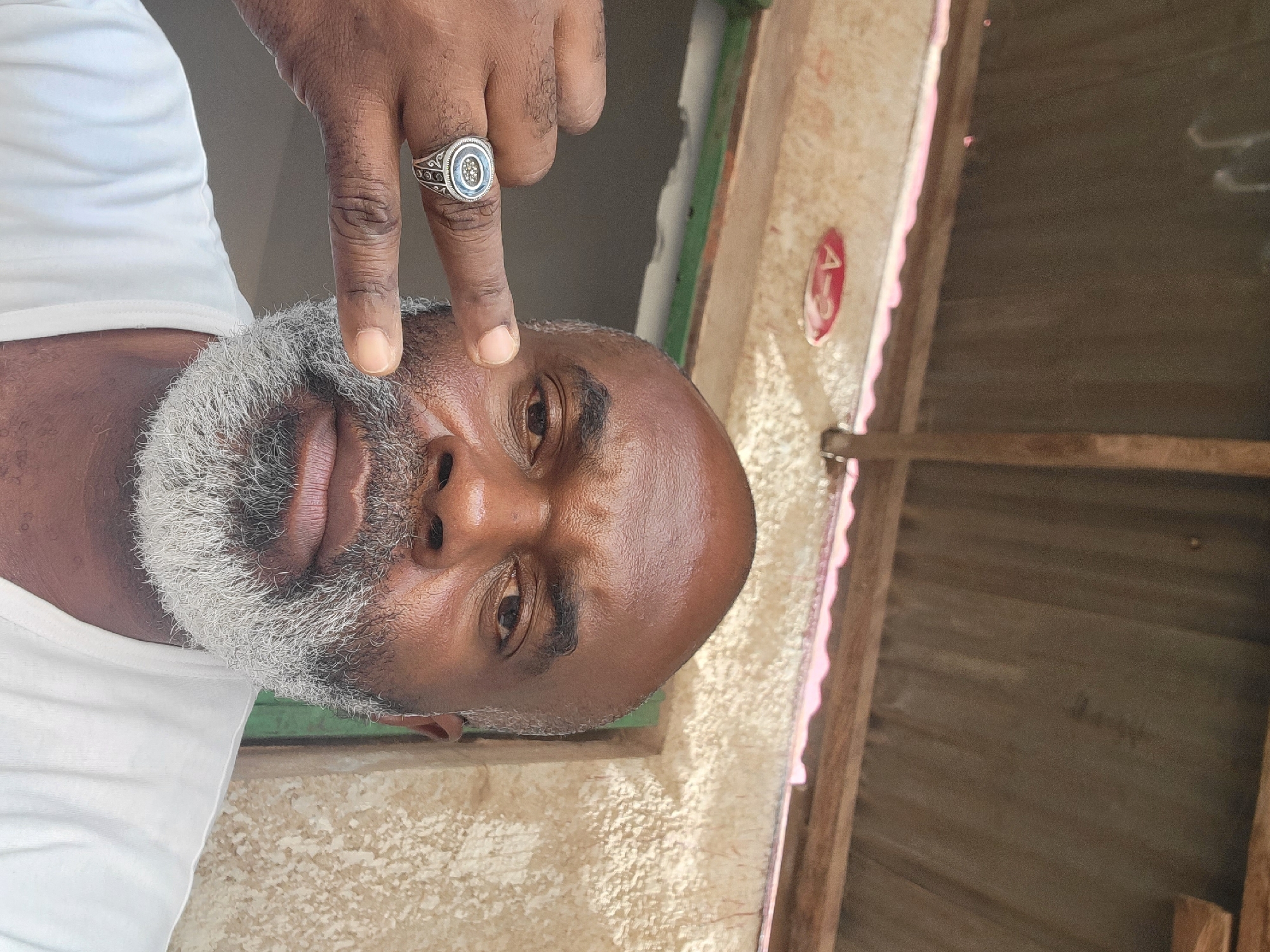 An upward-facing, medium close-up portrait shot features an adult Black man with a bald head and a full, salt-and-pepper grey beard and mustache. He is wearing a white t-shirt. His right hand is raised, with the index and middle fingers extended, pointing towards his left eye, and he wears a distinctive silver ring with a blue oval centerpiece and intricate filigree on his index finger. His expression is neutral.
The background reveals an indoor or semi-outdoor setting. To the left of the man is a light-colored, textured wall, possibly made of concrete or plaster. Behind him, a glimpse of a green-painted frame or window sill is visible. To the right, a wooden ceiling or structure with reddish-brown planks is present. A notable detail on the textured wall is a red, oval-shaped plaque with the white text "A-3" clearly visible. The lighting suggests daylight. The image was captured in Maiburiji, Nigeria.