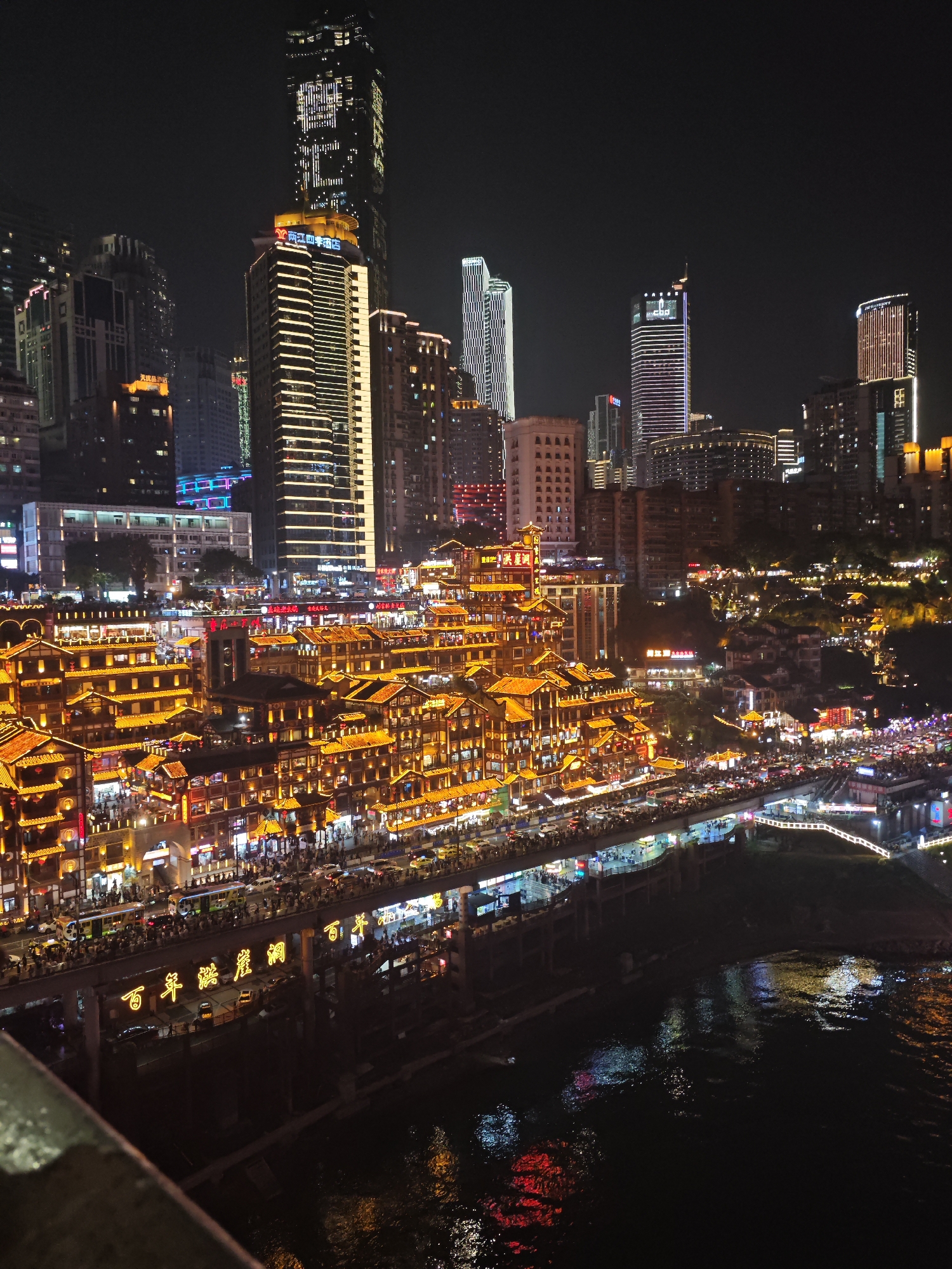 The image depicts a nighttime view of Hongyadong, a popular tourist attraction in Yuzhong District, Chongqing, China. The multi-story traditional stilt houses are heavily illuminated with warm golden lights, creating a vibrant scene. Pedestrians crowd the walkways along the structures. Behind Hongyadong, modern skyscrapers rise into the night sky, also lit up with various patterns and colors. A bridge spans a dark river in the foreground, with illuminated Chinese characters visible on its side. The river reflects the lights of the city. The sky is dark and devoid of stars.