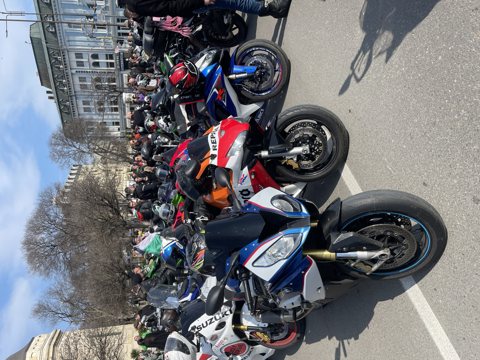 The scene depicts an outdoor gathering of numerous sport motorcycles and a crowd of people on a sunny day in Varna, Bulgaria, specifically located near Nezavisimost Square with the Varna Municipality building visible.
Multiple motorcycles, primarily sportbike models, are parked in close formation on asphalt pavement marked with white lines. In the foreground, a blue, white, and red BMW S1000RR motorcycle with carbon fiber detailing on its front is prominently featured. Adjacent to it are an orange, red, and white Honda CBR displaying "Repsol" and "HRC" branding, and a blue and white Suzuki motorcycle with "SUZUKI" text and pink rim tape on its wheels. Other motorcycles, including a white Suzuki with "Lucky Strike" branding, are visible further back. Various helmets are placed on motorcycle seats and handlebars.
A large congregation of adult individuals, mostly men, fills the area around and behind the motorcycles. Many are attired in motorcycle jackets and protective gear, while others wear casual clothing. Individuals are observed standing, conversing, and looking at the motorcycles. One person, seated on a motorcycle in the mid-ground, has a Bulgarian national flag draped over their back.
The background features distinctive urban architecture under a clear blue sky. On the left, a portion of the ornate, light-colored Varna Municipality building, characterized by a visible domed tower and classical architectural elements, is framed. To the right, a multi-story building with a light blue lower facade, numerous windows, and a dark roofline stands across what appears to be a street. Bare trees, indicative of late winter or early spring, are visible between the buildings. Strong sunlight casts distinct shadows across the pavement.