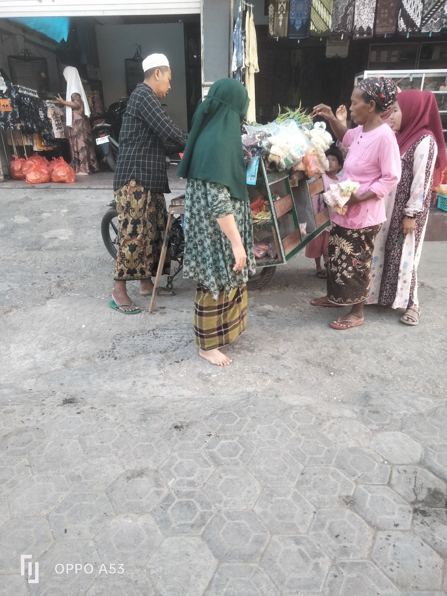 An outdoor street scene in Badung, Indonesia, features several individuals interacting near a marketplace. On the left, a man wearing a black and white checkered shirt and a white peci cap stands with a scooter. He is accompanied by a woman in a patterned brown and white dress and a white headscarf, who appears to be browsing items displayed in a store. In the center, a woman in a green headscarf and patterned dress stands facing a cart filled with various goods, including what appears to be produce and packaged items. To her right, a woman in a pink long-sleeved shirt and a patterned headscarf is interacting with the cart, holding a bundle of items. Behind her, a person wearing a red hijab is visible. The ground is paved with hexagonal concrete tiles. Red plastic bags are visible on the left side of the frame, near the store entrance. The overall lighting suggests it is daytime.