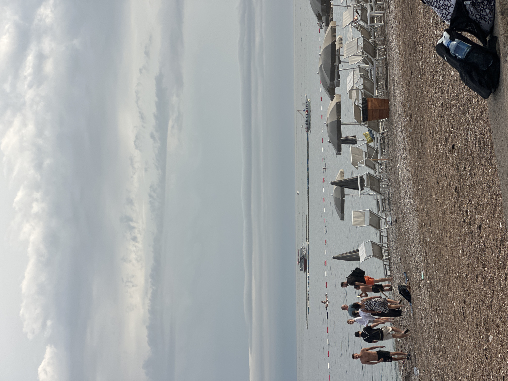 The image depicts a cloudy day at a pebbly beach in Budva, Montenegro. In the foreground, a group of people are gathered on the shore, some walking and others standing and talking. Beach chairs and umbrellas are set up in neat rows behind them, suggesting a designated beach area. The sea extends to the horizon, with a few boats visible in the distance. A rope with buoys separates the swimming area from further out. The overall atmosphere is relaxed, with people enjoying the beach despite the overcast weather.