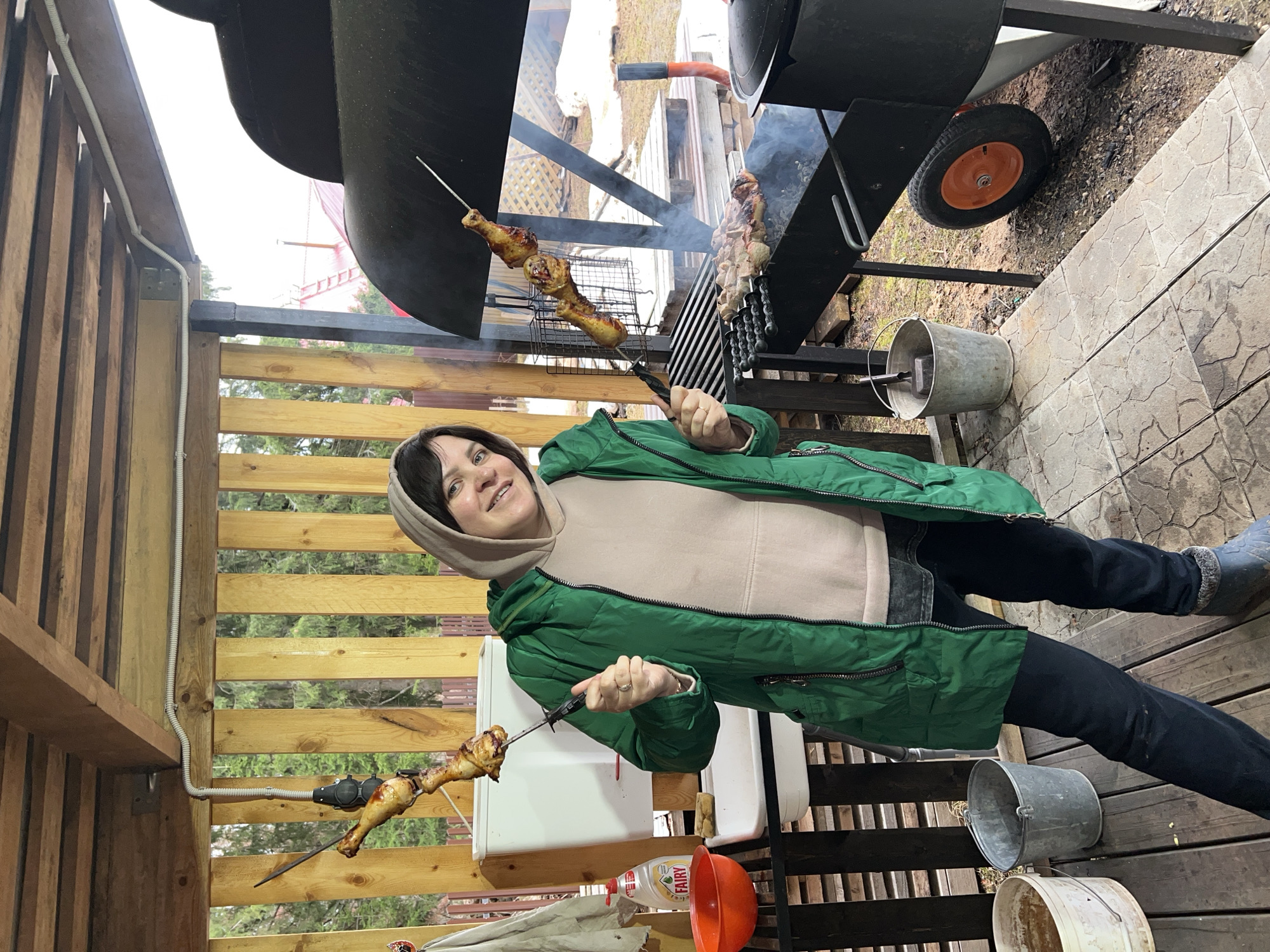 A woman in a green jacket and a light-colored hoodie is holding skewers of cooked chicken legs, standing on a wooden deck with a stone-paved area visible in the upper right corner of the image. She is near a barbecue grill with meat skewers cooking on it. In the background, there are wooden fences and trees. An orange bucket and bottles are visible near a white box on the deck. There are also a few metal buckets. The location is near Bol'shakino, Russia.