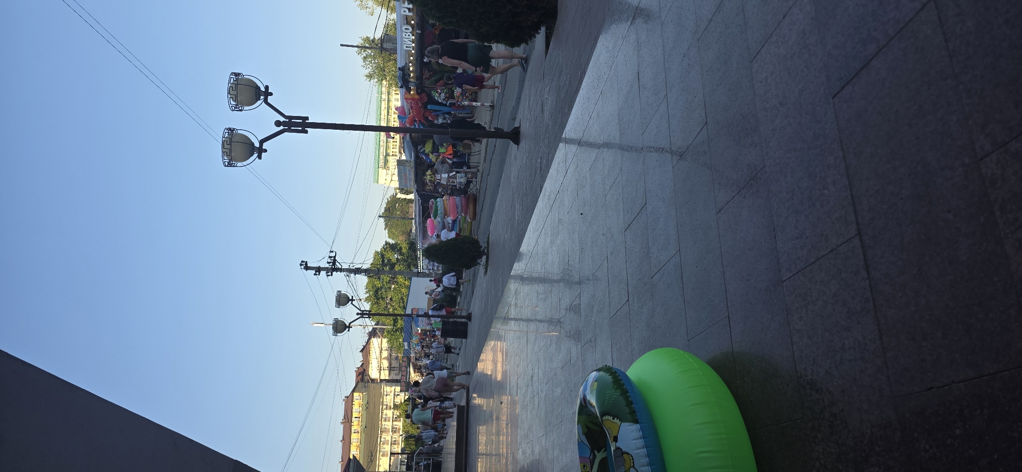 The image shows a scene on a street in Vityazevo, Russia, likely a resort town given the presence of beach accessories. The foreground features a blue and green inflatable pool ring. The middle ground depicts a paved walkway with reflections of the sky and overhead lights. A street lamp with two lights is prominent. Various market stalls are set up along the walkway, displaying items such as inflatable rings, balloons, and other merchandise. Several people are walking along the street, browsing the stalls or moving past. In the background, buildings with multi-colored facades suggest a commercial or residential area. The sky is clear and blue, indicating a sunny day. The overall atmosphere appears to be that of a bustling street scene in a tourist destination.