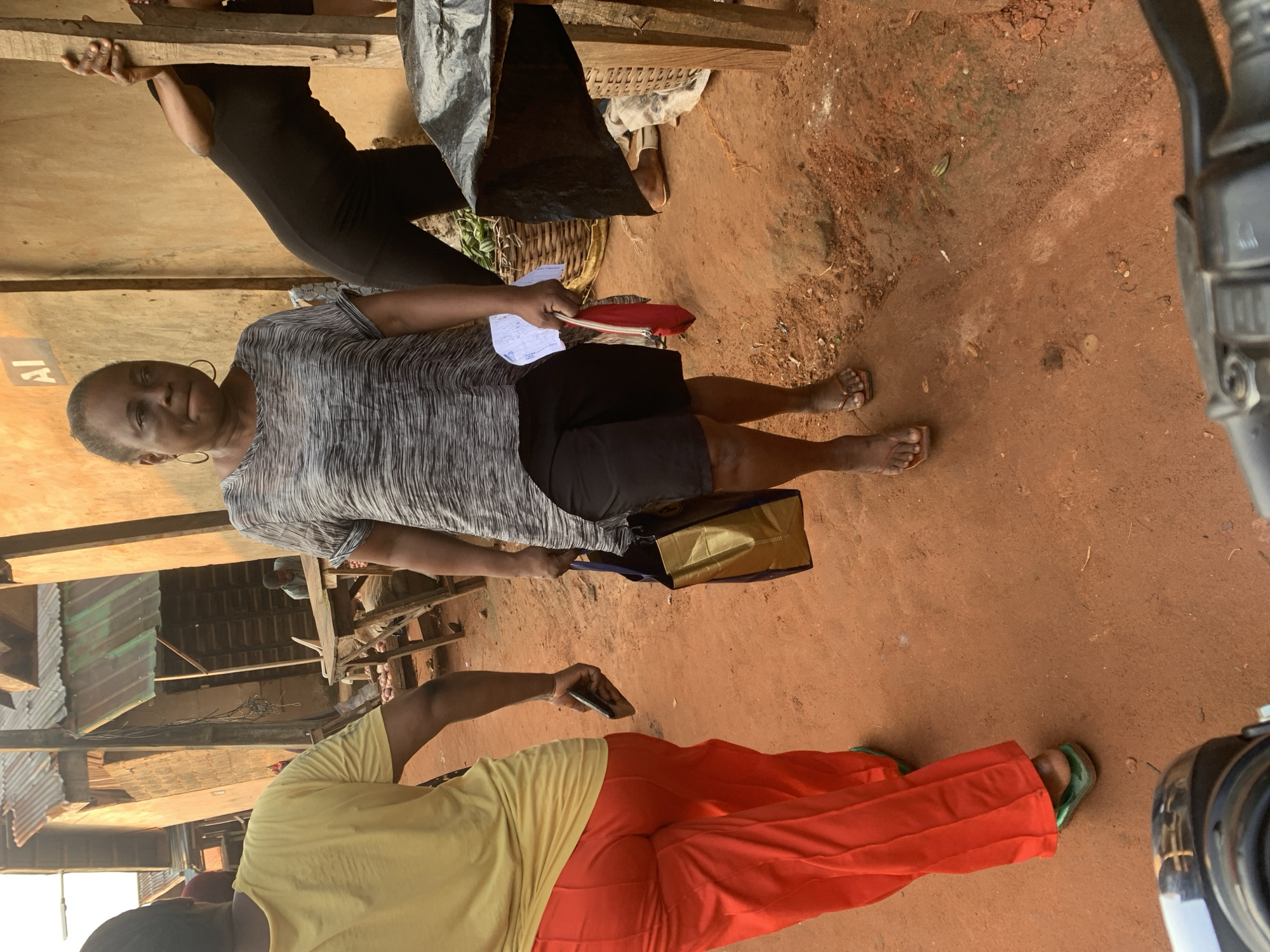 This image depicts a scene in a marketplace in Shagamu, Nigeria. In the foreground, a person wearing bright orange pants and green flip-flops is walking away from the camera. To their right, a woman in a grey and black patterned shirt and black shorts is walking towards the camera, carrying a red clutch and a gold and blue tote bag. She is holding a piece of paper. Further in the background, another person in black is leaning against a wooden post, and a man is visible behind a table with produce. The environment appears to be an outdoor market with stalls and buildings constructed with corrugated metal roofs and earth-toned walls. The ground is covered in reddish-brown dirt. The lighting suggests it is daytime, possibly late afternoon given the warm tone of the light.