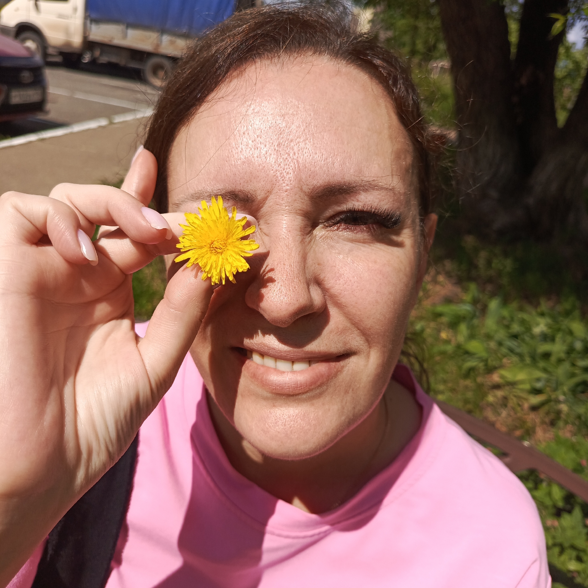 A woman is holding a dandelion in front of her eye. She is wearing a pink t-shirt and is outdoors, with trees and greenery in the background. A truck and a car are visible in the distance, suggesting an urban or suburban setting. The image is a close-up of the woman's face and hand. The sunlight suggests it is daytime.