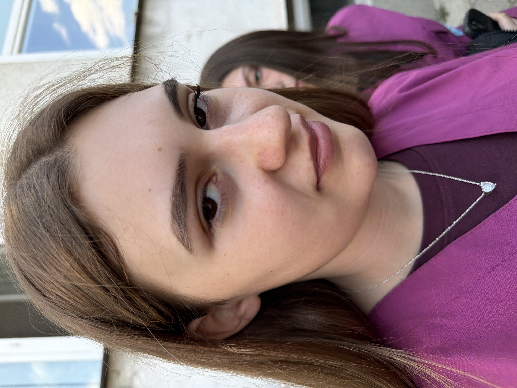 A close-up, angled shot features a young woman with fair skin, brown hair, and brown eyes. She is wearing a purple shirt and a delicate silver necklace with a heart-shaped pendant. In the background, another woman with long dark hair is partially visible, also wearing a purple shirt. The background suggests an outdoor setting with a building facade visible. The image provides no direct indication of being taken in Panagyurishte, Bulgaria.