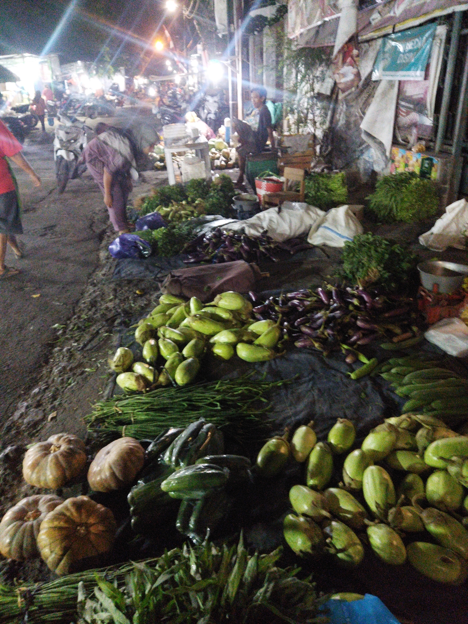 The image depicts an outdoor market scene at night, possibly in Semarang, Indonesia, based on the urban setting and night market atmosphere. The ground is dark and appears to be a dirt or paved surface. In the foreground, various fresh produce is laid out on dark tarps and on the ground. There are several large, ridged pumpkins, a pile of green gourds or squashes, bundles of long green beans or similar vegetables, and a large quantity of purple eggplants. Behind the produce, a person in a light purple garment is seen carrying a load on their back, walking away from the camera. In the background, several motorcycles are parked, and there are bright lights illuminating the scene, suggesting a bustling night market. People are visible in the background, though their details are indistinct due to the lighting and distance. Stalls or structures with various goods are also present in the background.