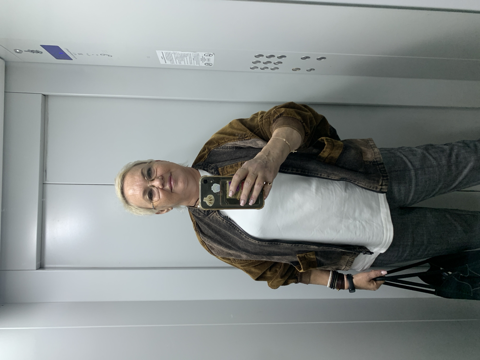 A blonde woman with glasses is standing in an elevator, taking a mirror selfie. She is wearing a white t-shirt, a brown corduroy jacket, and grey pants. She has several bracelets on her left wrist and is carrying a black bag. The elevator walls are metallic grey, and there are buttons on the right wall. The image appears to have been taken in Chaikovsky, Russia.