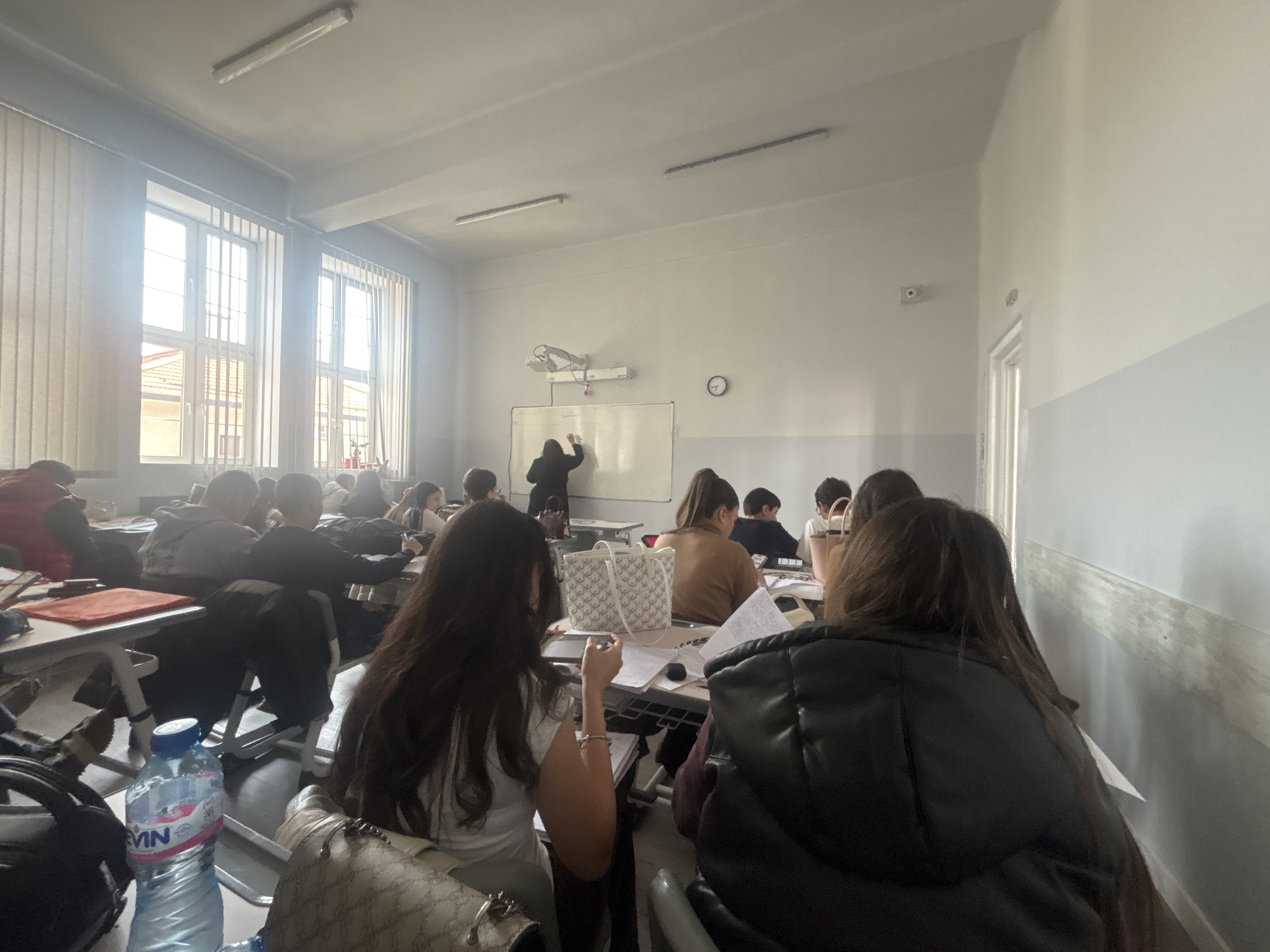 The image shows a classroom in Sofia, Bulgaria. The room is filled with students sitting at desks, facing a whiteboard where a person is writing. Light enters through large windows. There are desks, chairs, papers, and personal items like bags and water bottles. The environment is a typical school setting with standard lighting and painted walls.