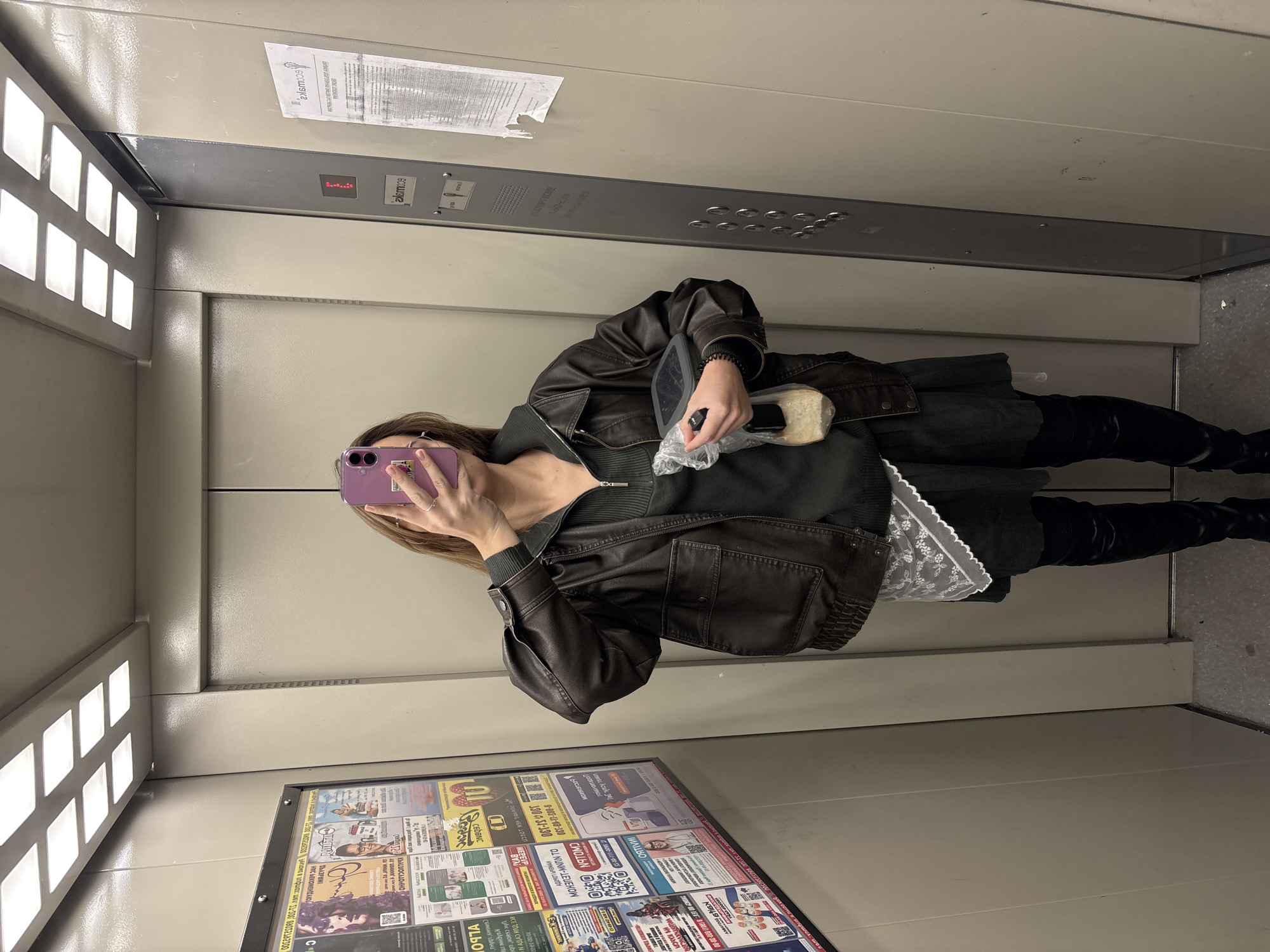 A woman is taking a selfie in what appears to be an elevator. She is holding a purple smartphone in front of her face, obscuring her features. She is wearing a dark jacket over a dark top, and black leggings or boots. In her other hand, she holds a container with food, possibly a sandwich.
The elevator's interior is utilitarian, with grey metal walls and bright fluorescent lights. A notice or advertisement board with various posters and text in Russian is affixed to the wall near the floor. Some of the Russian text on the board refers to what the OCR identifies as a phone number: 8-805-11-10-130, and mention of “Ust-Ilimsk”, which may indicate a location in that city in Russia. Another poster mentions “coronatomo” . An elevator control panel is above her head. The overall scene is compact and enclosed.
