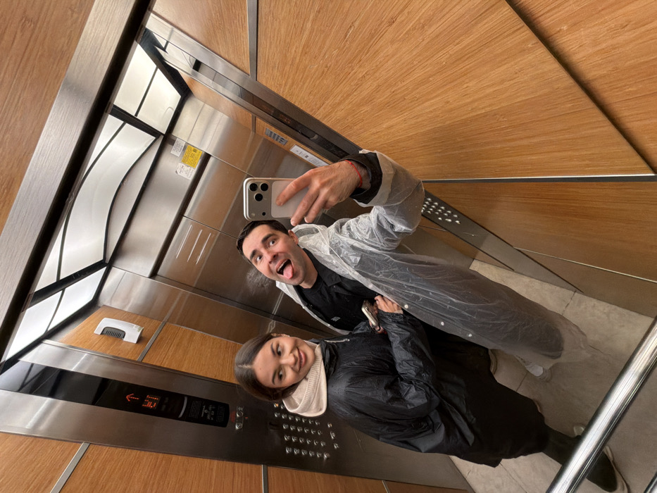 Inside a wooden-paneled elevator, a man in a clear raincoat holds a smartphone, taking a selfie. He sticks his tongue out at the camera. A woman in a dark jacket smiles beside him. The elevator's interior is a mix of metal and wood, with a panel showing an arrow pointing left, next to the number 30. Sunlight streams through the elevator shaft, reflecting off the metal surfaces.