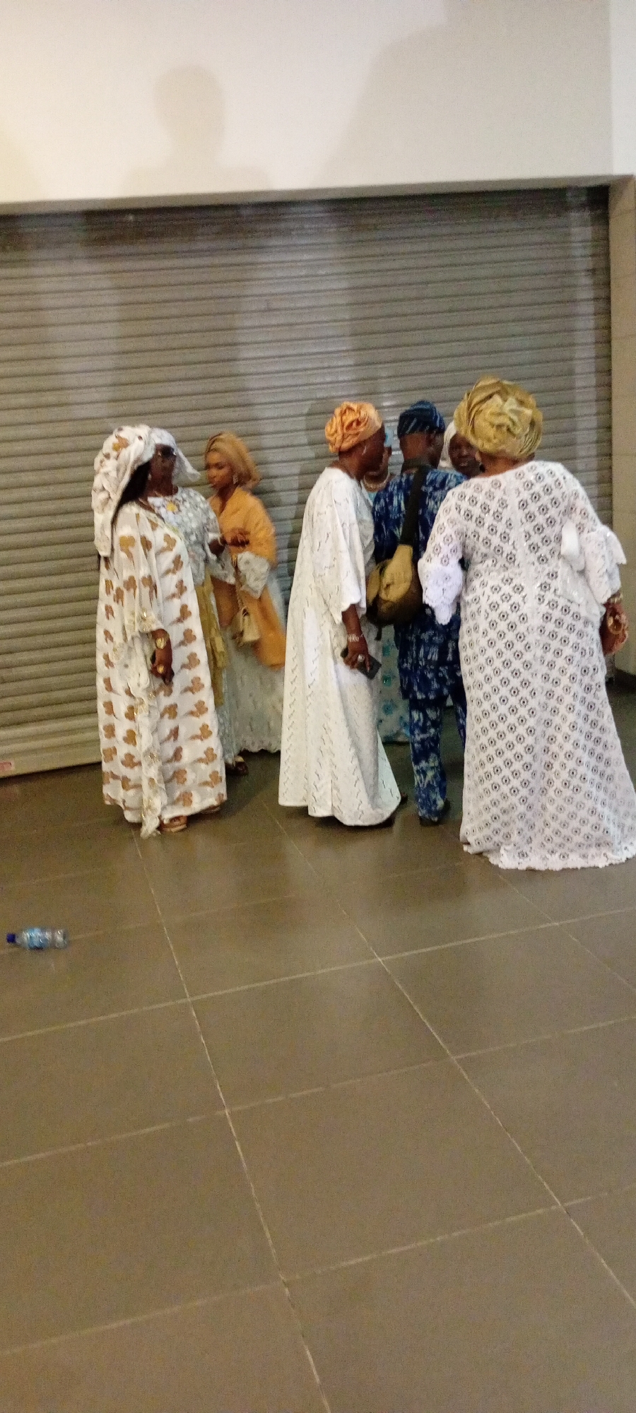 A group of five people, four women and one man, are standing in front of a large, corrugated metal roll-up door in what appears to be an indoor public space. The floor is tiled with large, square, tan-colored tiles. A plastic water bottle is lying on the floor in the lower left corner.
The women are all wearing traditional Nigerian attire with elaborate headwraps and long, flowing dresses. The woman on the far left is wearing a white and gold patterned dress and headwrap. Next to her, a woman in an orange headwrap and matching outfit is looking at her. To their right, a woman in a white dress and orange headwrap is facing away from the camera, speaking to the man. The man is wearing a blue and white patterned outfit, a headwrap, and a bag slung across his shoulder. The woman on the far right is wearing a white lace dress and a golden headwrap, facing away from the camera.
The lighting suggests the scene might be indoors with artificial light or late in the day. The overall impression is of a gathering or event. The location is identified as Gandu Albasa, Nigeria.