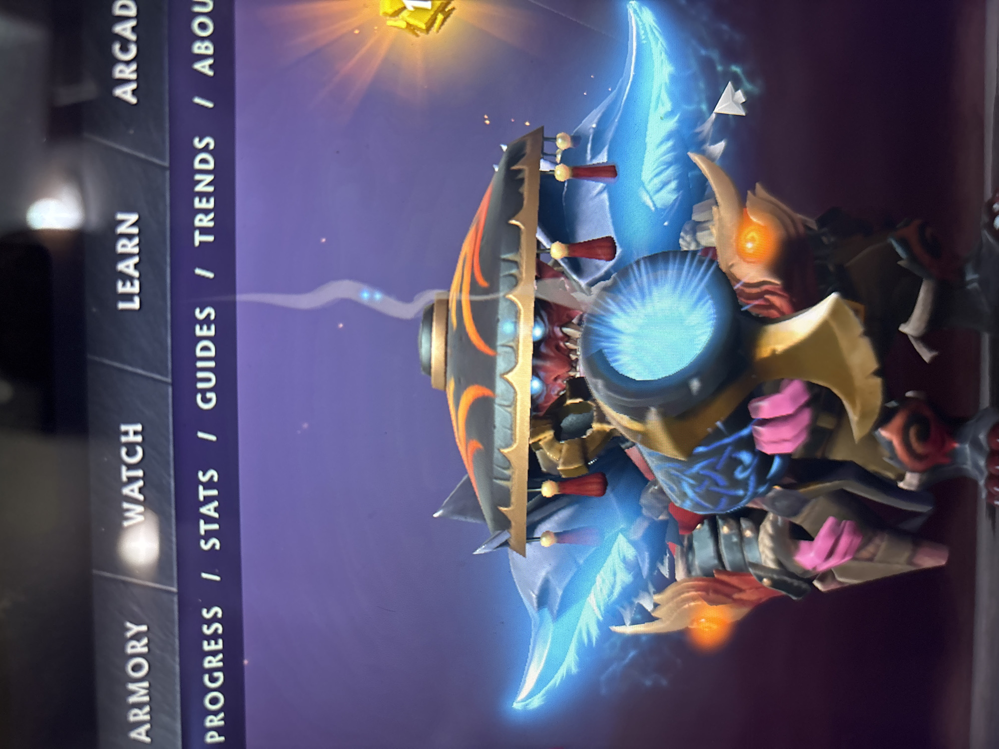 The image captures a static display of a video game interface on a screen. The central subject is an elaborately designed 3D character, likely a hero or monster, depicted against a dark purple and blue digital background with glowing particles. The character features a wide, flat, dark hat-like headpiece adorned with orange and black patterns and red tassels. Beneath this, a reddish, possibly demonic face with glowing orange eyes is visible. Prominent glowing blue energy elements, resembling wings or fins, extend from the character's sides, and a bright blue orb or device is centered on its front. The character also wears gold-toned armor and has pink/purple accents on what appear to be its hands or claws. In the upper right portion of the game display, a bright yellow, blocky icon, possibly representing gold or a reward, emits a strong light.
On the left side of the screen, a vertical navigation bar is visible, featuring white, capitalized text on a dark gray background. The main menu options displayed are "ARMORY," "WATCH," "LEARN," and "ARCADE." Adjacent to these, a secondary navigation area lists sub-categories separated by slashes: "PROGRESS / STATS / GUIDES / TRENDS / ABOUT." The overall scene presents a detailed character model within a game's user interface, suggesting a menu or character inspection screen.