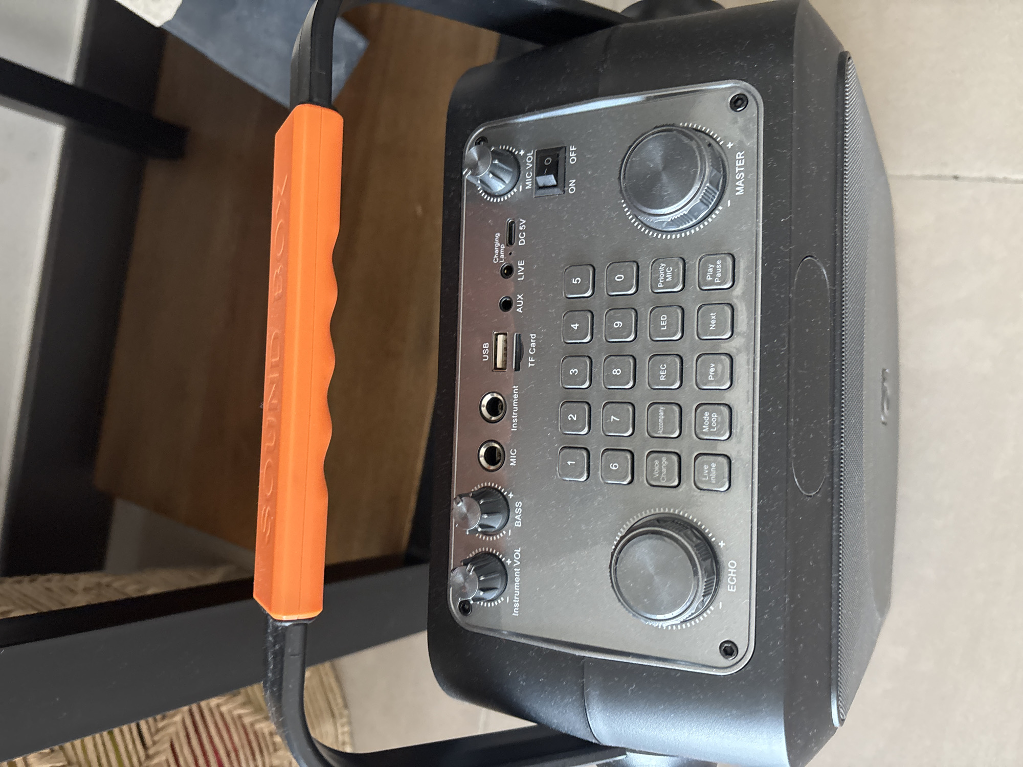 The image shows a close-up of the top panel of a portable speaker system, likely a karaoke machine or a similar audio device. The device is black with a metallic gray control panel. Prominently featured are various knobs and buttons, including "Instrument VOL," "BASS," "MIC," "Instrument," "TF Card," "AUX," "LIVE," "DC 5V," "MIC VOL," and an "ON/OFF" switch. Below these are a numerical keypad (0-9) and several function buttons such as "Voice Change," "Accompaniment," "REC," "LED," "Priority MIC," "Prev," "Next," "Play/Pause," and "Mode." There are also two large rotary knobs labeled "ECHO" and "MASTER." A bright orange handle with the embossed text "SOUND BOX" is visible at the top. The speaker itself is at the bottom of the frame, with a mesh grill. The overall scene is dusty, suggesting the device may be in a less-than-pristine environment. The background is blurred but appears to show furniture, possibly a wooden table and chairs, with a woven basket or textured material to the left. The lighting suggests an indoor setting. There are no people visible in the image, and no specific geographical markers indicate a location in Dubai, United Arab Emirates.
