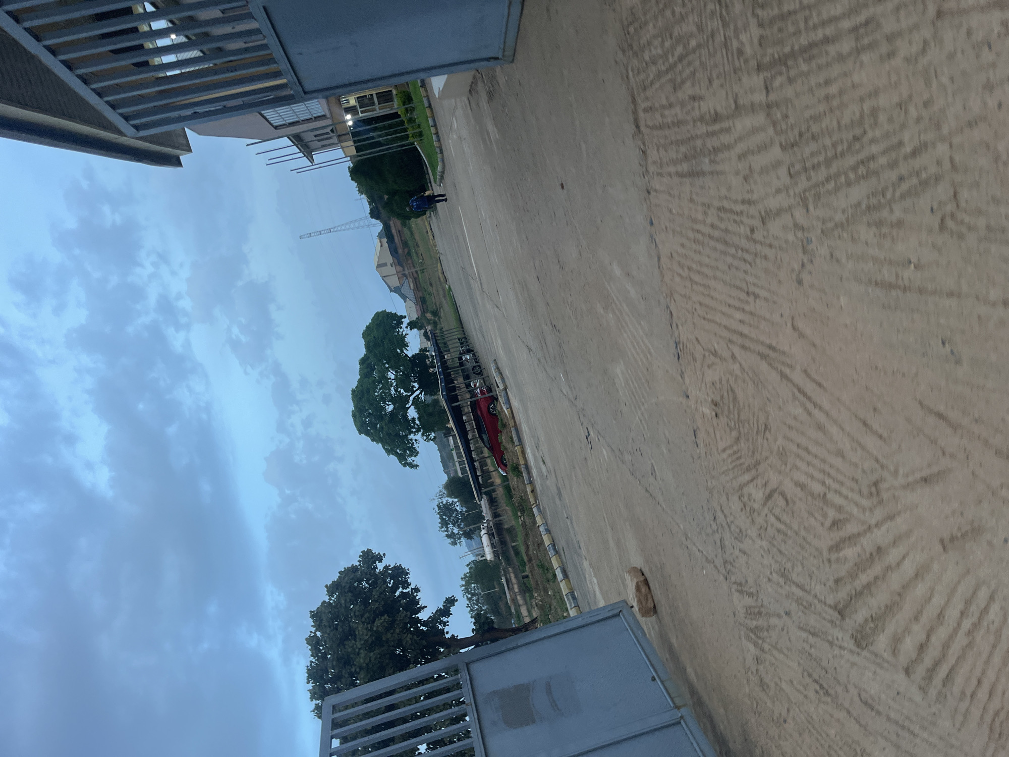 The image depicts an outdoor scene at dusk, possibly in Gura Top, Nigeria. The foreground is dominated by a paved area with visible tire tracks and a large, grey metal gate partially open on the left. To the right, another section of a grey metal fence is visible, leading towards a building with a sloped roof. The building has several windows and appears to be a commercial or institutional structure.
In the middle ground, a red car is parked under a covered structure. Further into the background, there are trees, some with dense foliage, and a tall transmission tower can be seen against the cloudy sky. A single individual, dressed in dark clothing, is walking away from the viewer on the paved area. The sky is overcast with a mix of blue and grey clouds, suggesting impending or recent rain. The overall impression is of an entrance to a compound or facility.