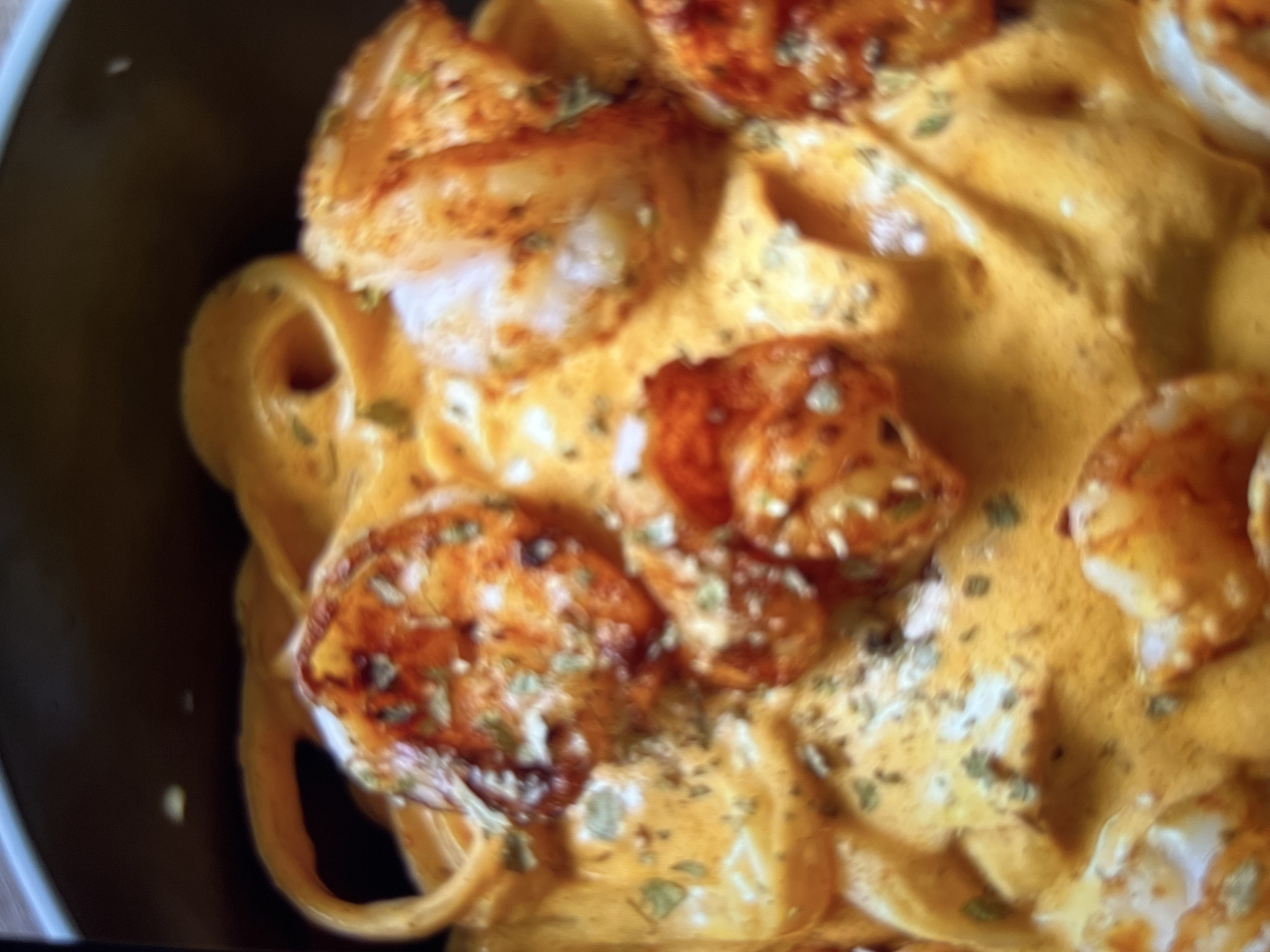A close-up, slightly blurry overhead shot shows a bowl of pasta with shrimp in a creamy orange sauce. The pasta appears to be linguine, with several strands visible and coiled. The shrimp are pink and appear to be sautéed, with some seasoning sprinkled on top. The sauce is rich and thick, coating the pasta and shrimp. The overall impression is a decadent and flavorful dish. The location in the city of Abuja, Nigeria is not discernible from the image.