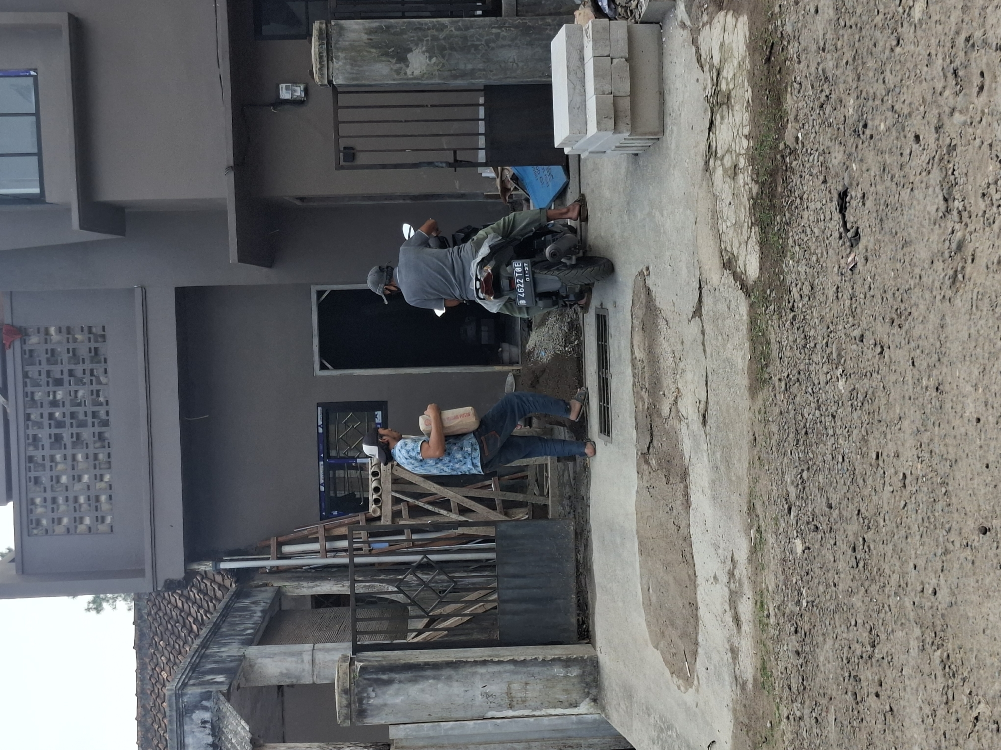 The image depicts an outdoor scene in the city of Ciheulang Tonggoh, Indonesia, featuring a construction site adjacent to a two-story building. In the foreground, a man wearing a patterned blue shirt, jeans, and sandals walks from left to right, carrying a bag of cement. To his right, another man wearing a gray t-shirt and pants, with a baseball cap, is seated on a motorcycle facing away from the camera. The motorcycle is parked on a concrete surface.
Behind the individuals, a partially constructed building with dark gray walls is visible. A ladder leans against the building. Several stacks of concrete blocks are present near the motorcycle. A metal gate with a diamond pattern is located to the left of the scene, partially obscuring a covered porch with a tiled roof. The ground surface transitions from concrete to a rough, gravelly area in the foreground. The overall environment suggests ongoing construction or renovation work.