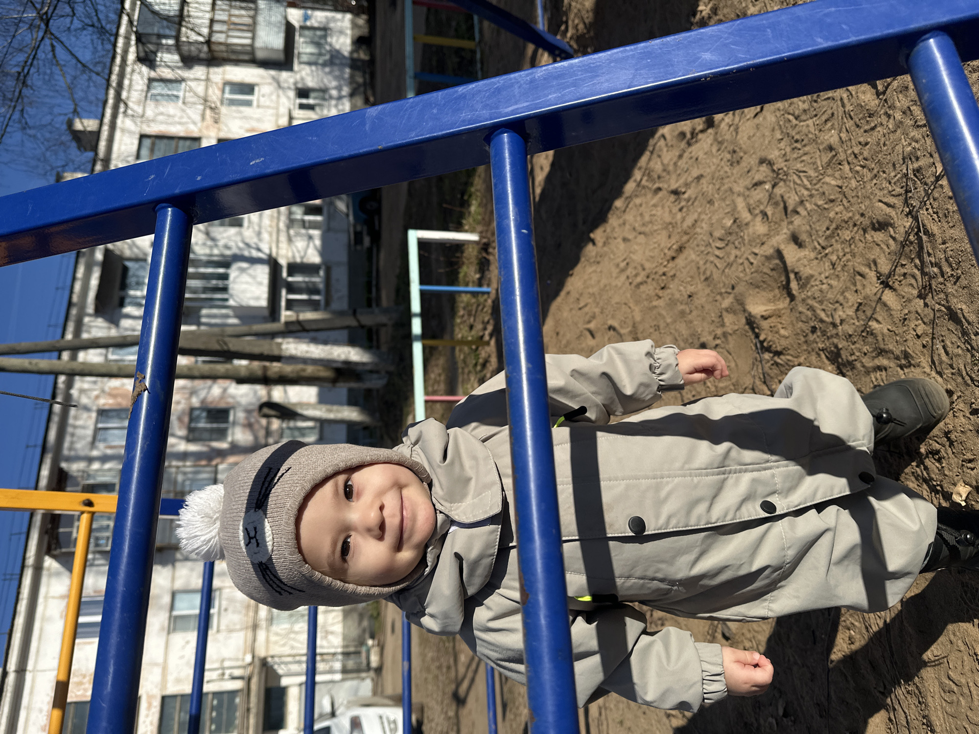 A young child, likely a toddler, is captured smiling brightly directly at the camera on a playground, with the scene set in Chaikovsky, Russia. The child is dressed in a light grey full-body snowsuit or romper, fastened with black buttons, and a matching light brown beanie hat featuring a white pom-pom and subtle embroidered cat-like eyes.
The child is positioned in the middle ground, framed by the bright blue metal bars of a playground structure in the foreground, creating a dynamic perspective. The ground below is a sandy or dirt surface, exhibiting visible footprints and some dry vegetation. Strong, distinct shadows are cast across the ground by the playground equipment, indicating bright, clear daylight.
In the background, a multi-story residential building with numerous windows is visible, partially obscured by bare tree branches silhouetted against a clear blue sky. Another section of playground equipment, including yellow and blue bars, can be seen behind the child to the left. The time of day appears to be sunny daytime, suggesting pleasant weather for outdoor play, possibly early spring or late autumn given the bare trees and warm clothing. The child's happy, smiling expression conveys a sense of joy and contentment. No visible text is present in the image.