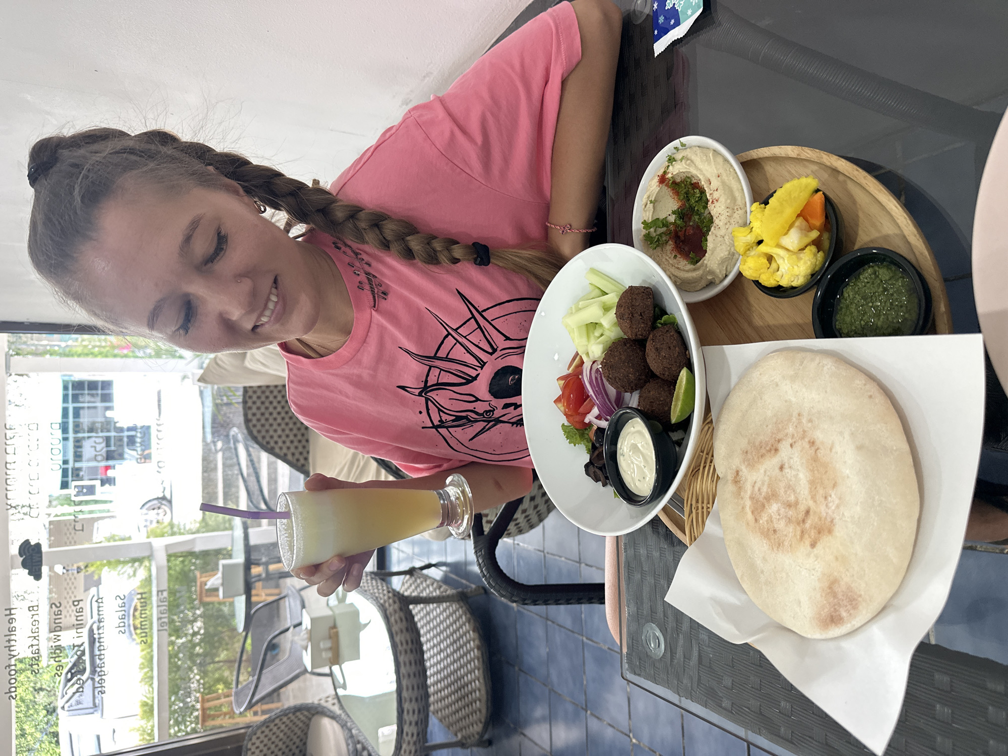 A young woman with a braided blonde ponytail, wearing a pink t-shirt with a graphic design, sits at a table in a restaurant in Baan Mae Nam, Thailand, holding a glass of yellow juice with a purple straw. In front of her is a meal consisting of falafel, a salad with tomatoes, onions, cucumbers, and olives, a bowl of hummus, a bowl of pickled vegetables, a side of green sauce, and a piece of pita bread on a white paper liner. The background shows a window with text on it, chairs, and tables. The table is dark and glossy.
