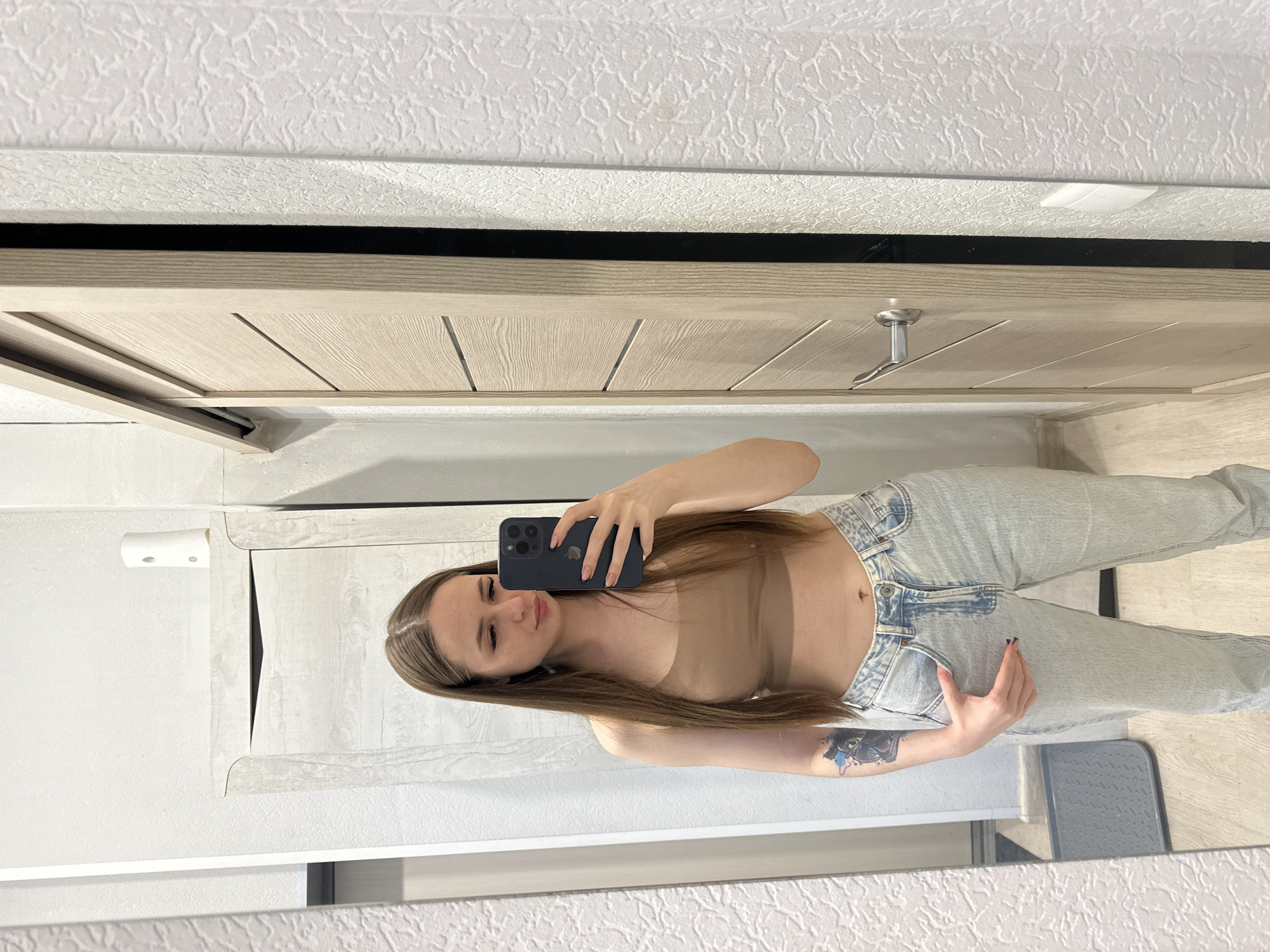 A woman stands in front of a mirror, holding a smartphone to take a selfie. She is wearing a nude-colored top and light-wash jeans. A tattoo is visible on her left arm. The background includes a wooden door, a white wall with texture, and a white cabinet. The location is inside a room, presumably in a private residence.