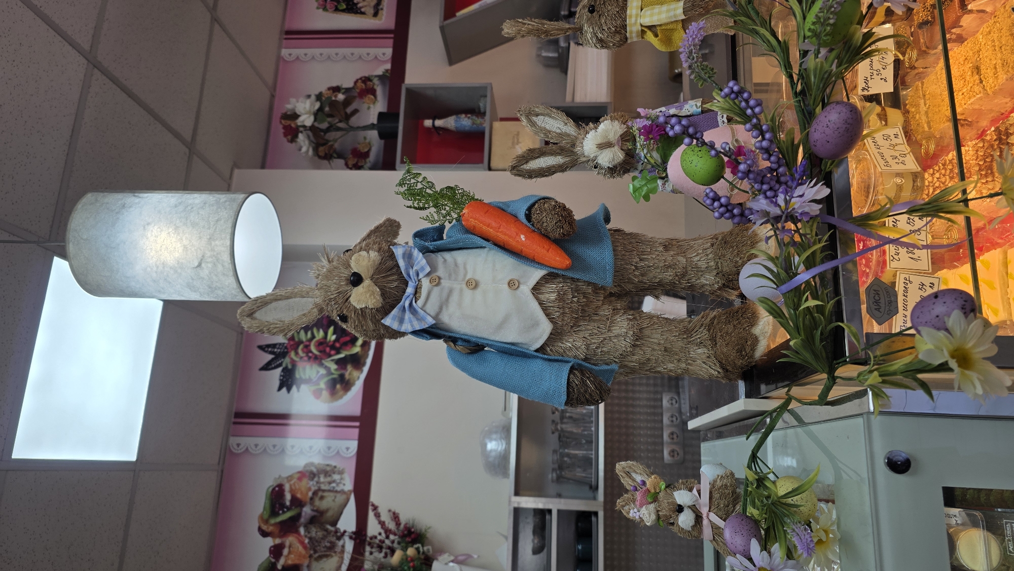 Here is a description of the image:
The image displays a decorative arrangement likely found inside a retail or commercial establishment, possibly in Blagoevgrad, Bulgaria. The focus is on Easter-themed decorations, particularly straw bunny figures dressed in clothing and holding carrots, positioned among floral arrangements and decorative Easter eggs. The scene is inside, adjacent to a display case filled with pastries or desserts, identifiable by the signage written in Bulgarian.
The environment includes shelving units and tiled walls, suggesting a store or bakery interior. A contemporary cylindrical lamp fixture is visible in the background, as are posters or advertisements featuring pastries. The overall impression is a festive and commercially oriented display.