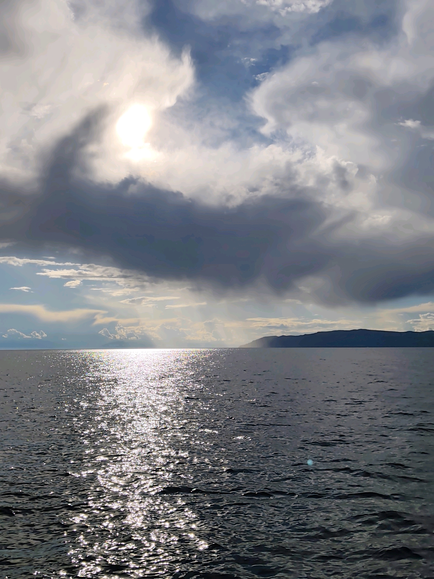 The image depicts a body of water, likely Lake Baikal, with a distant, dark, mountainous shoreline. The sky is dramatic, with heavy, dark clouds partially obscuring a bright sun. Rays of sunlight break through the clouds, creating a shimmering path across the water's surface. The water itself is dark and choppy, reflecting the light in numerous bright sparkles. No people or vessels are visible in the scene. The general atmosphere is one of natural grandeur and possibly impending weather change due to the dramatic cloud formations.
