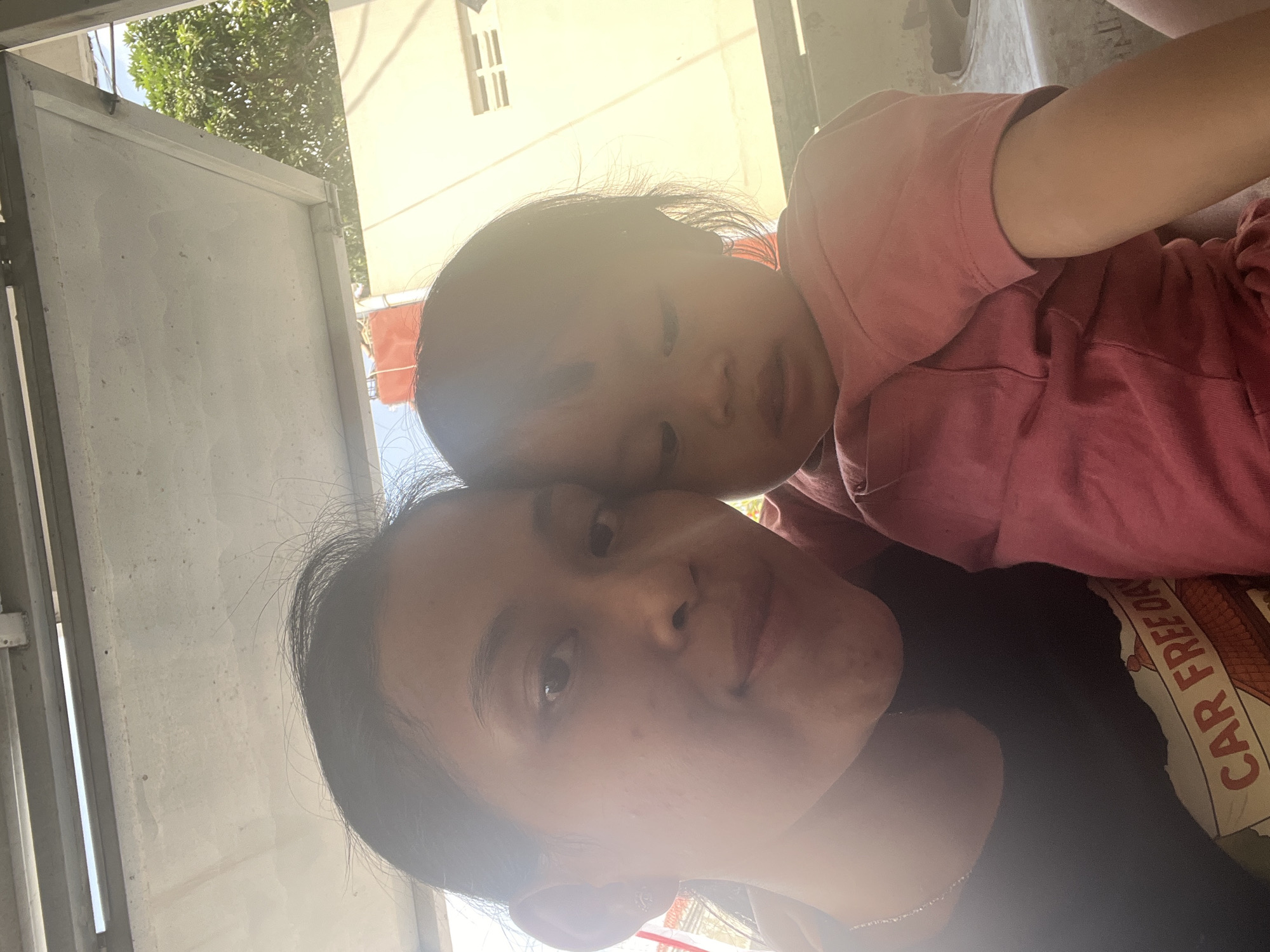 A selfie captures a woman and a young child, likely mother and daughter, in an outdoor setting. The woman, on the left, is looking at the camera. She has dark hair and is wearing a black t-shirt with a design that appears to say "CAR FREE DAY". The child, on the right, is looking down. They are both wearing pink shirts. The background suggests an urban or suburban environment, with a building and trees visible. The lighting indicates it is daytime, with bright sunlight creating some lens flare. The presence of a metal structure overhead suggests they might be under a covered patio or carport. The scene appears to be within Kecamatan Pati, Indonesia, based on the provided location information.
