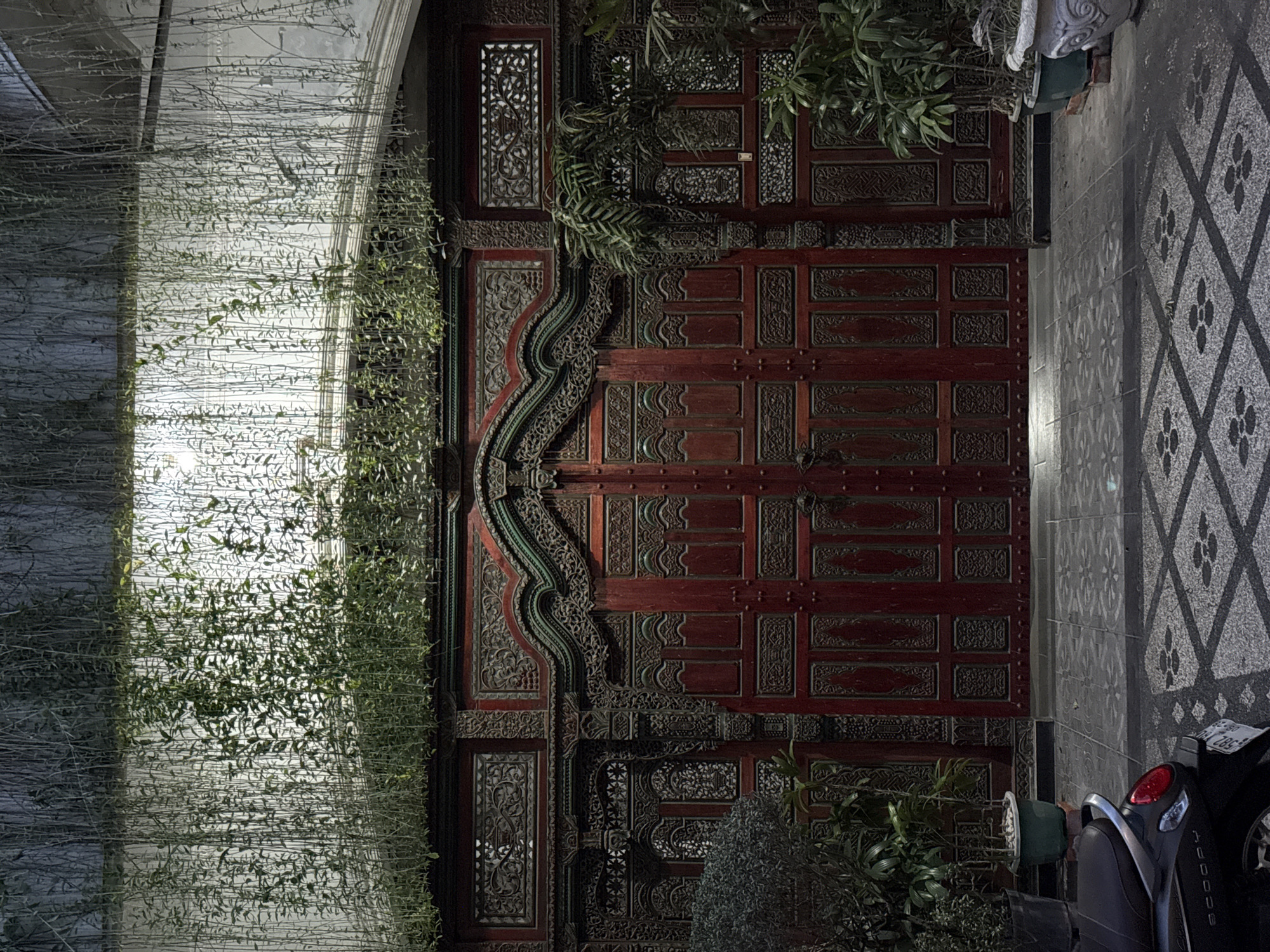 The image captures a doorway in Kerobokan Kelod, Indonesia. The ornate, dark-red, wooden double door is the central focus. The door features intricate carvings and panels. Draping greenery, resembling vines or hanging plants, cascades down the left side of the door and onto the adjacent wall. The wall is white and textured. The ground to the right of the door is paved with a patterned tile. A motorcycle is parked on the right side of the frame. The scene is illuminated with artificial light, creating highlights and shadows.