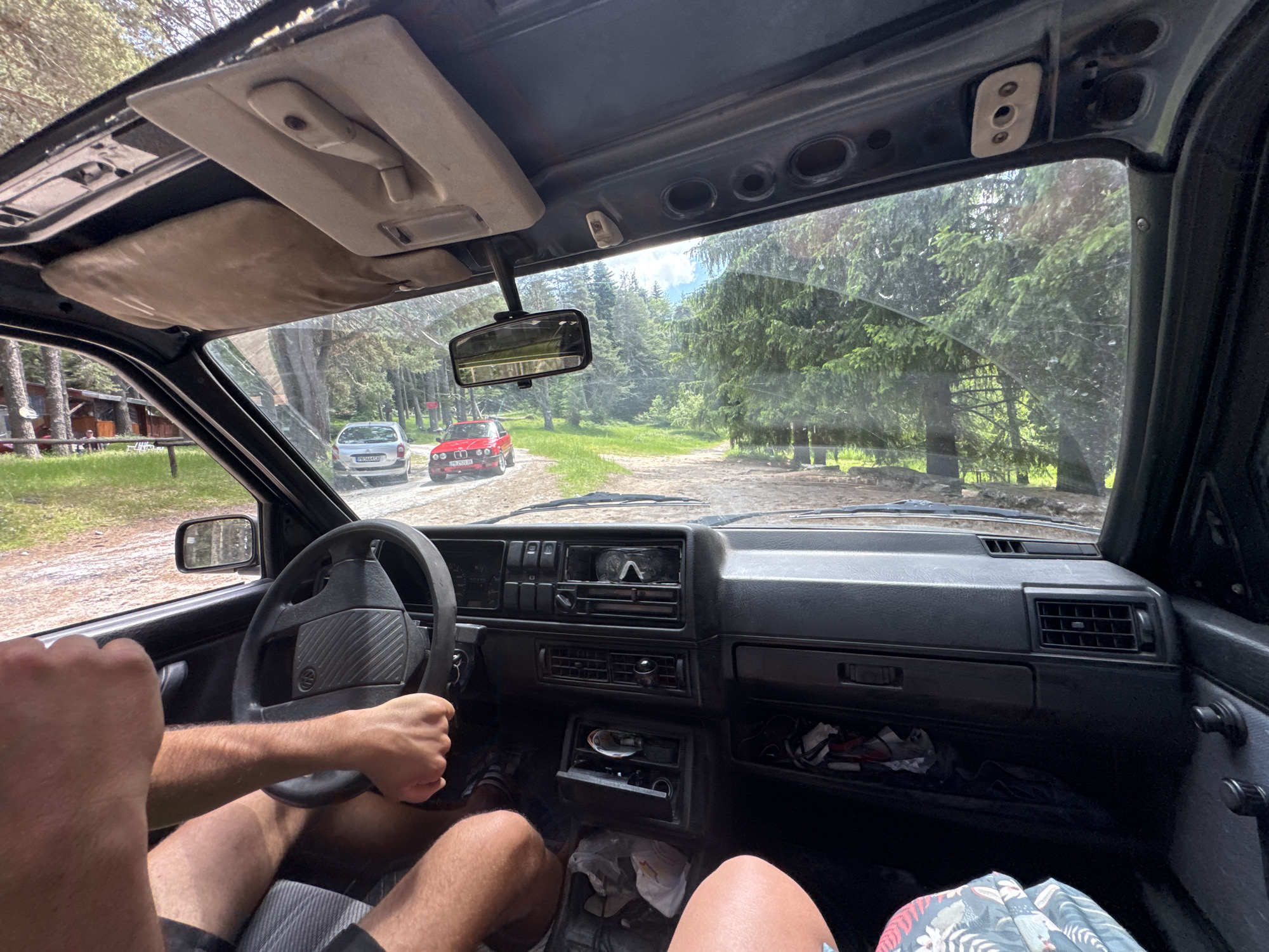 The image is taken from the interior of a car, looking forward through the windshield. The car appears to be driving on a dirt road surrounded by a dense forest of pine trees. In the distance, another car, a red hatchback, is visible on the road ahead, with a silver hatchback behind it. The car's interior shows a dashboard with an air conditioning vent, a radio, and a steering wheel. The driver's hands are visible on the steering wheel. The overall scene suggests a scenic drive through a rural or natural landscape. There is no indication of the city of Dedovo, Bulgaria.