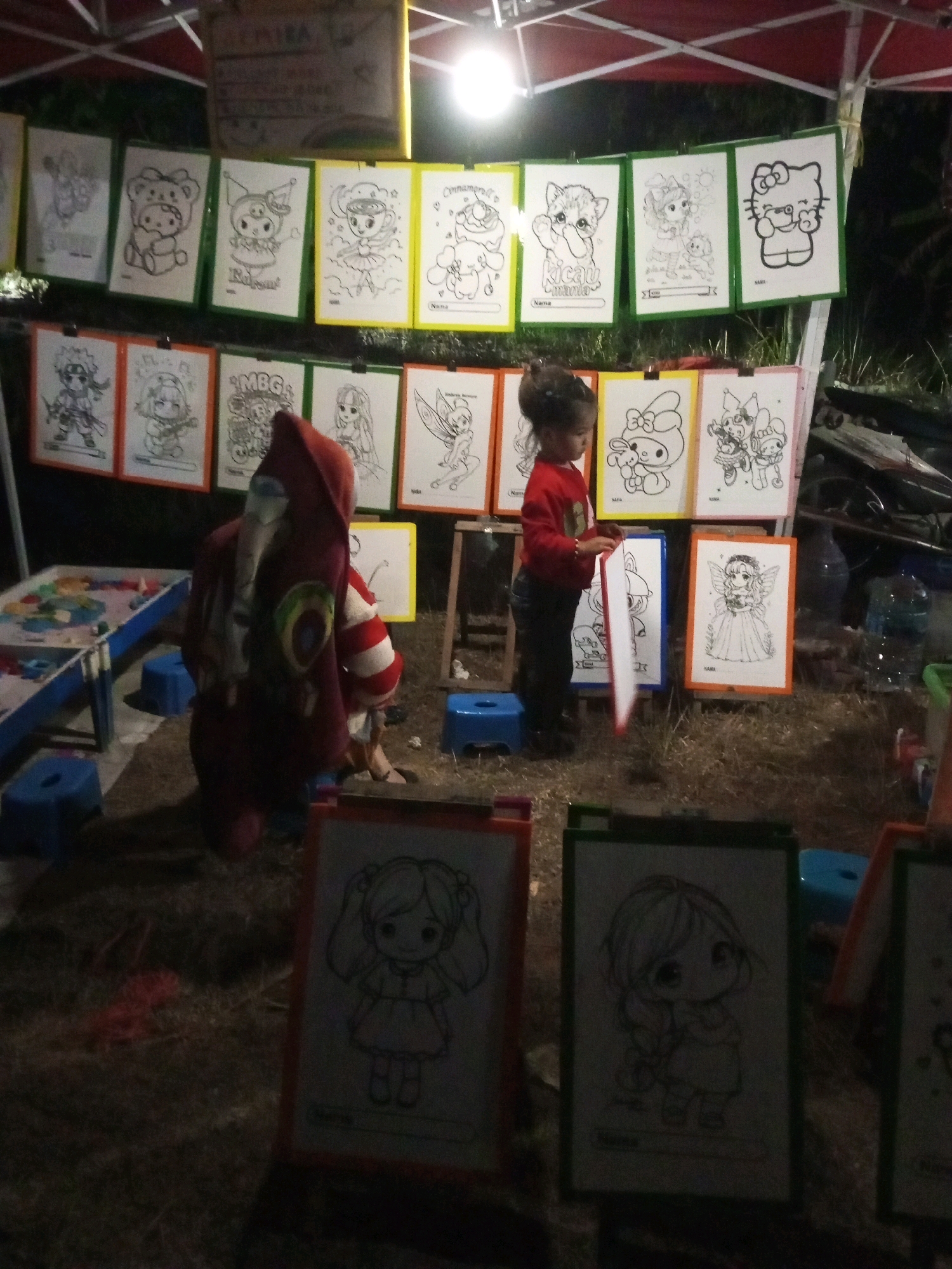 The image depicts an outdoor scene at night, possibly a market stall, in Ngadikerso, Indonesia. A young child, dressed in a red hooded garment with striped sleeves, is kneeling and holding a white sheet of paper. The child is facing away from the camera. Behind the child, a display of coloring pages is hung on a green and yellow frame. These pages feature various cartoon characters, including Hello Kitty, a bear-like character, and other illustrated figures. To the left of the child, a blue table with a colorful surface, possibly a craft or activity table, is visible, along with blue plastic stools. In the foreground, two framed coloring pages are propped up on the ground, one featuring a character with pigtails and the other with braided hair. The ground is covered with dry grass and dirt, and a red string or rope is visible on the ground near the table. The overall lighting suggests artificial illumination from overhead lamps, casting shadows and creating a dimly lit atmosphere.