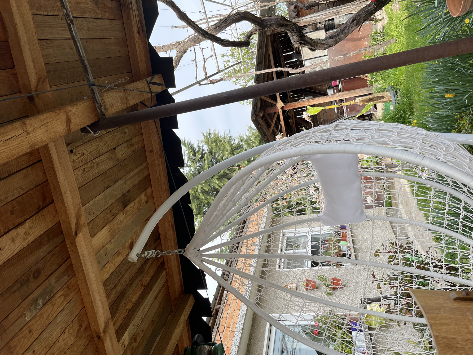 A slightly tilted, wide-angle shot displays a hanging white wicker chair with a white cushion, suspended beneath wooden eaves and a brown metal support bar in Karavelovo, Bulgaria. The chair hangs outside a cream-colored building with a window, brick detailing, and potted plants. Lush greenery and trees fill the background. Overhead, bare tree branches intertwine with a decaying wooden structure. The wooden eaves of a nearby building are visible on the left.