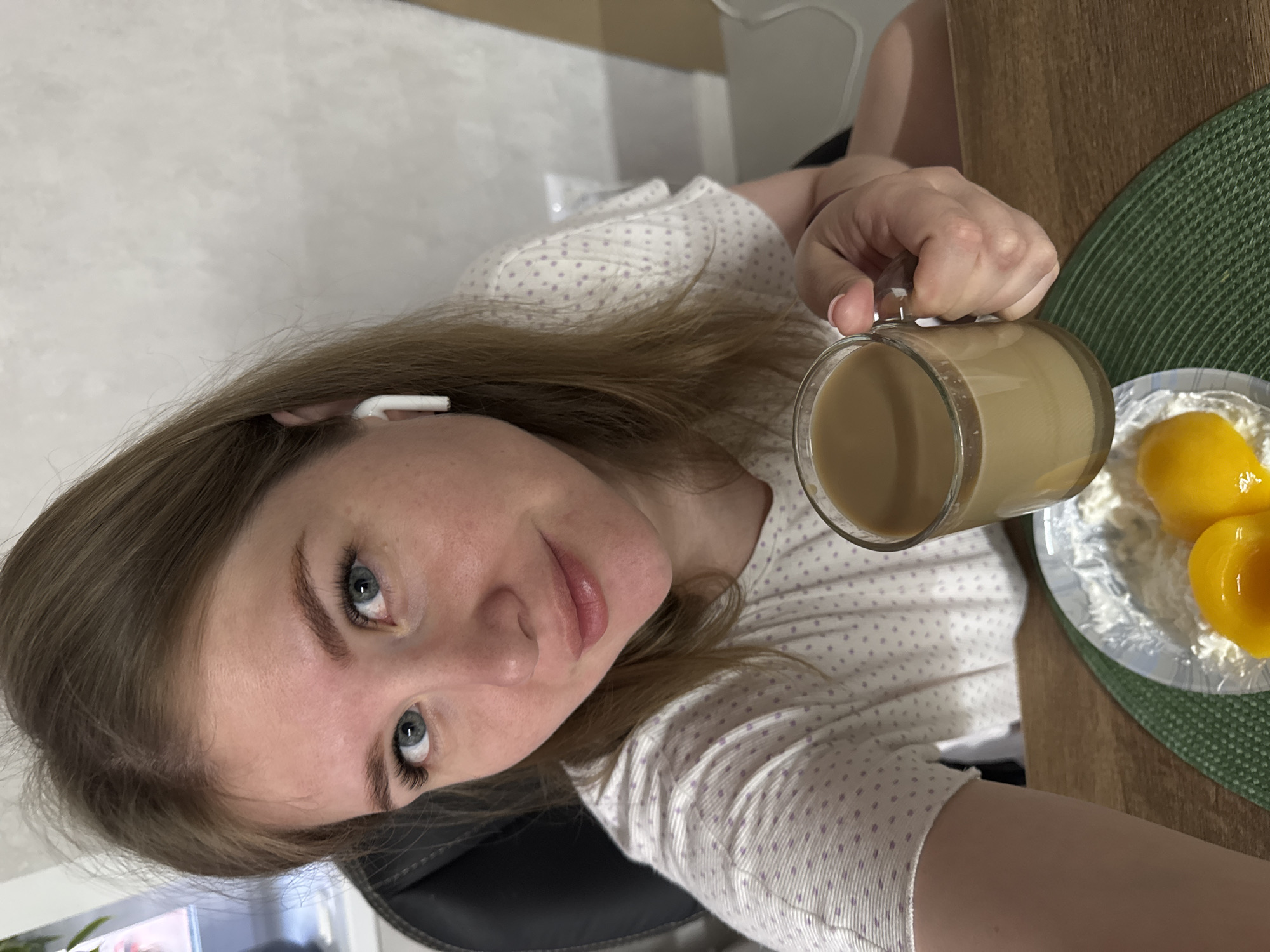A woman with light brown hair and blue eyes holds a glass mug filled with a tan liquid, likely coffee with milk. She is wearing a white shirt with small purple polka dots and a white wireless earbud in her left ear. To her right is a small green circular placemat with a white plate holding three halved peaches. The background appears to be an interior space, possibly a kitchen or dining area, with light-colored walls. The image is taken from a slightly above and angled perspective. There's not any specific information in the image to confirm it is in the city of Gornyi (Dvurechenskoe S/P), Russia.