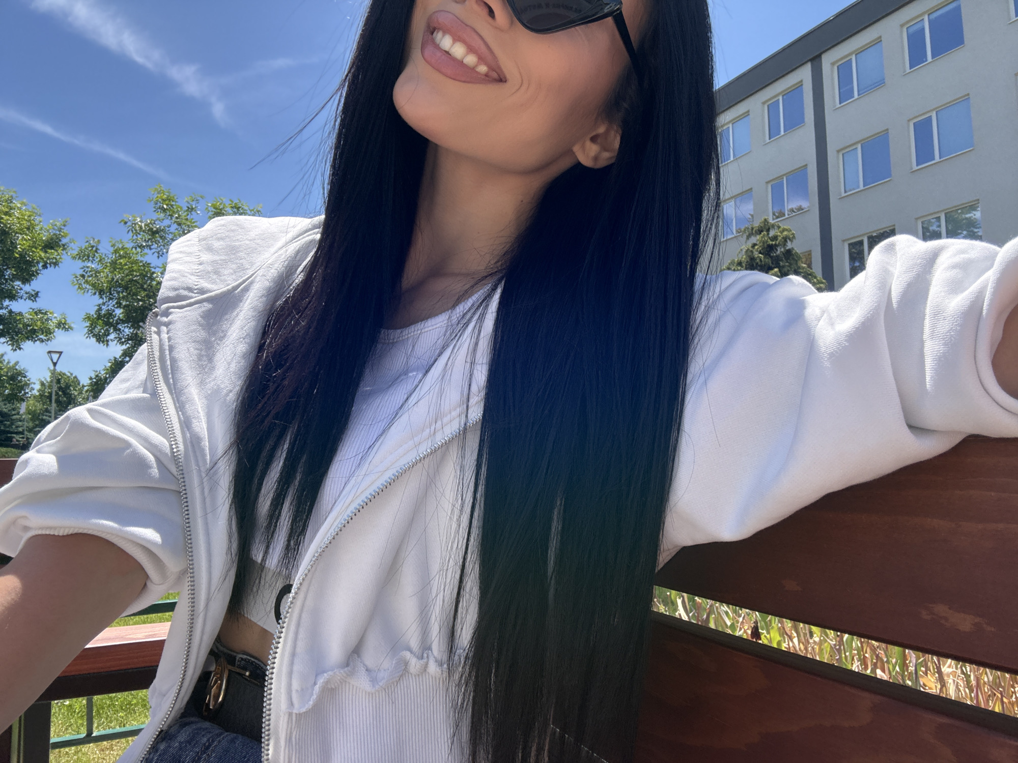 A woman with long black hair and sunglasses is sitting on a bench outdoors, smiling. She is wearing a white jacket and a crop top. The background shows trees, a building, and a blue sky with some clouds. The bench appears to be made of wood.