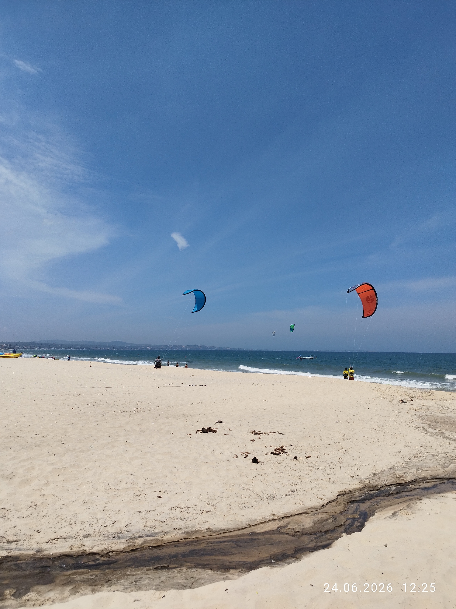 This image depicts a bright, sunny day at Mui Ne beach in Phu Thuy Ward, Vietnam. The foreground is dominated by a wide expanse of light-colored sand, marked by a dark, narrow channel of water flowing from right to left. Scattered debris, such as twigs and dried leaves, are visible on the sand.
In the midground, the turquoise ocean meets the sandy shore with gentle waves. Several individuals are present on the beach and in the water. Two prominent kites are visible: a large, bright blue kite soaring in the air over the water, and an orange kite with black and white accents, also airborne, with two people standing on the beach below it. Further out at sea, other kites and a small boat can be observed, suggesting kite surfing or kite boarding activities.
In the background, a hazy coastline with distant hills and structures is visible under a clear blue sky with scattered wispy clouds. A yellow structure, possibly a boat or kayak, is on the far left of the beach. The overall scene conveys a relaxed, recreational atmosphere characteristic of a popular beach destination. A timestamp, "24.06.2026 12:25", is present in the bottom right corner of the image.
