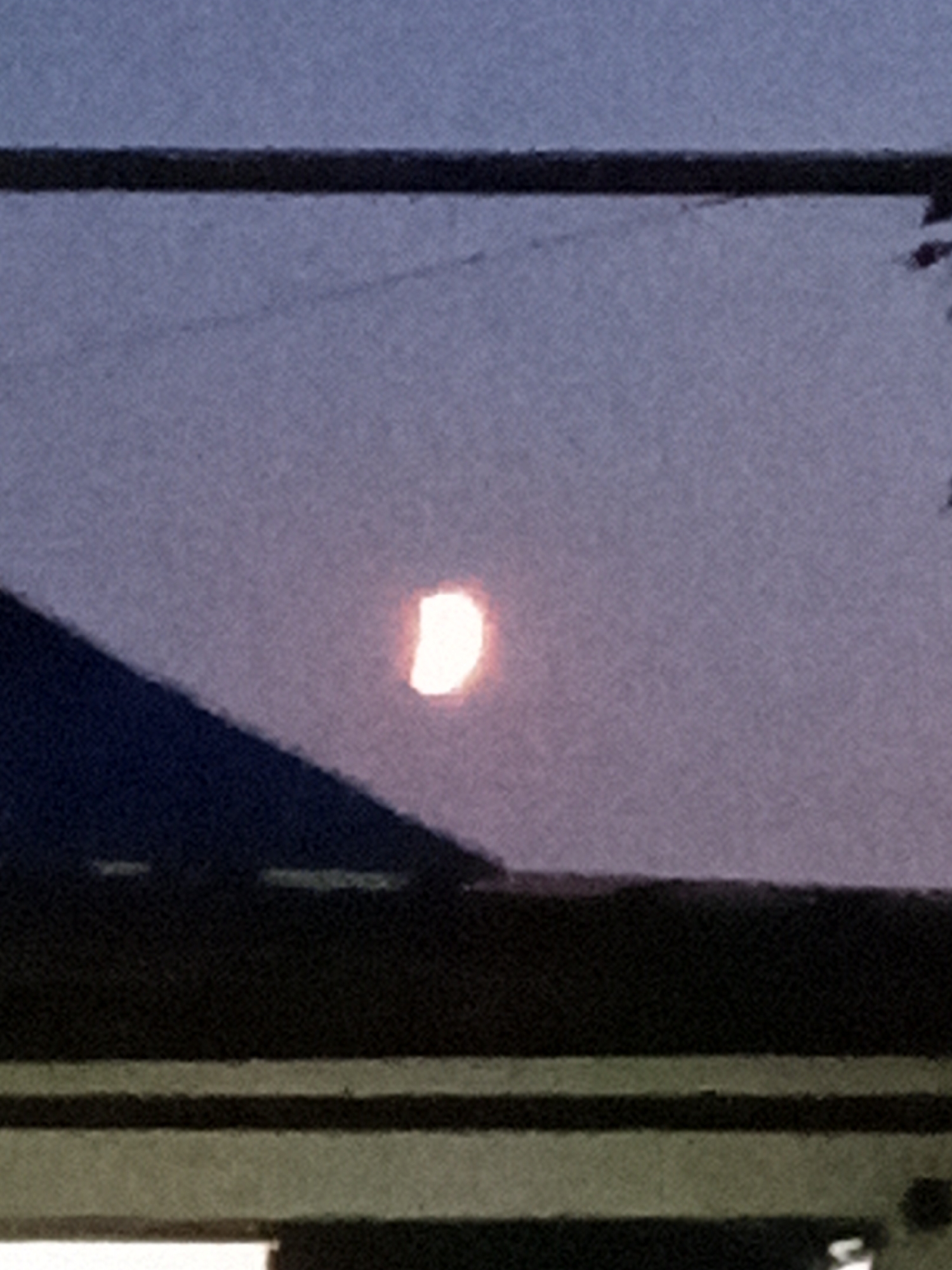 The image shows a partially eclipsed sun or moon in a twilight sky. The celestial body is bright white with a curved edge. Below the sky is a dark, angled structure that appears to be a roof or the side of a building, with horizontal lines of lighter color beneath it, suggesting multiple levels or structures. The overall scene is dim and slightly hazy. There are no people or discernible objects in the image. The location of Rudny, Kazakhstan, is not visually represented in the image.
