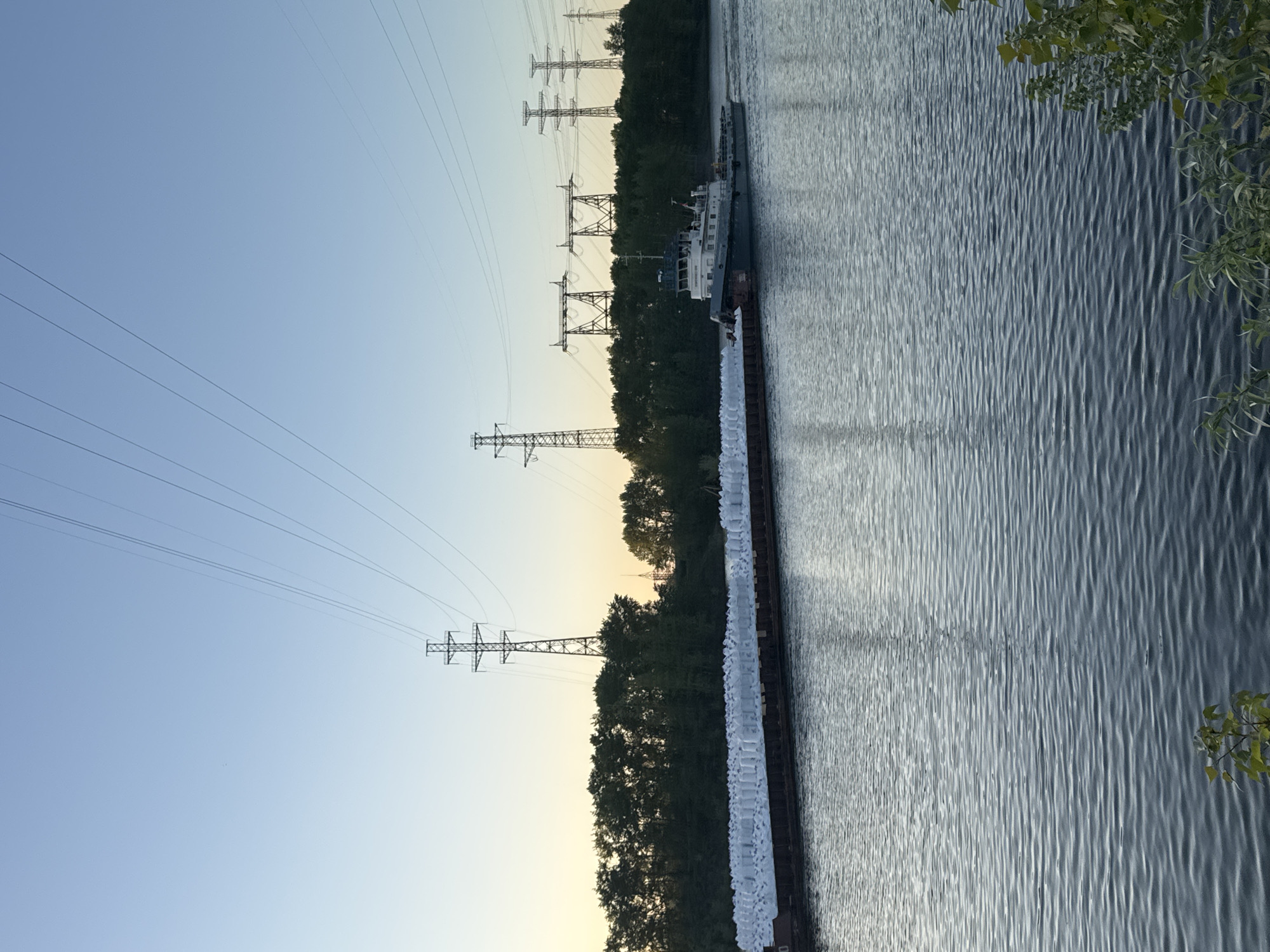 The image depicts a view of the Kama River near the city of Chaikovsky, Russia. A barge is navigating the waterway, with a tugboat attached to its side. The barge is loaded with what appear to be stacked white bags or packages. Transmission towers and power lines are visible along the shoreline, running adjacent to a line of trees. The sky is a light blue, and the water shows wave patterns.