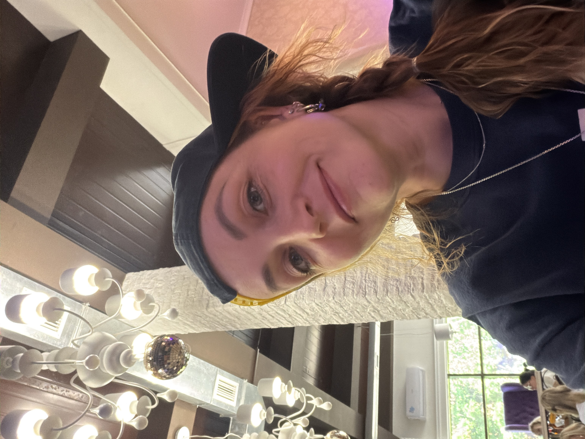 A selfie shows a person wearing a dark hooded sweatshirt and a black baseball cap looking down at the camera. The person has light skin, brown hair, and is wearing earrings and a necklace. The background includes a chandelier with multiple lights, a white ceiling with dark wood beams, a window with green trees visible outside, and a hand sanitizer dispenser on a white wall. In the reflection from one of the chandelier’s reflective lights is a seated person with dark hair, and a purple chair. The location is plausibly Chaikovsky, Russia based on the architectural elements and decor.