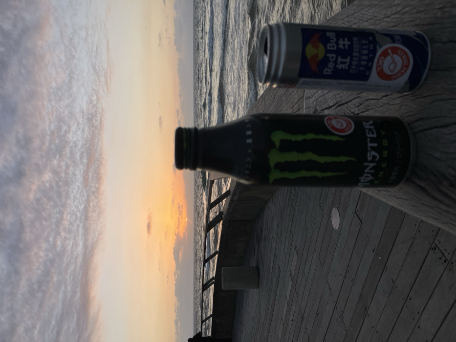 A bottle of Monster Energy and a can of Red Bull sit atop a weathered wooden railing, likely near a beach. The sunset paints the sky with hues of orange, pink, and purple, visible in the distance beyond the railing and the ocean. The scene is peaceful, with a calming view.