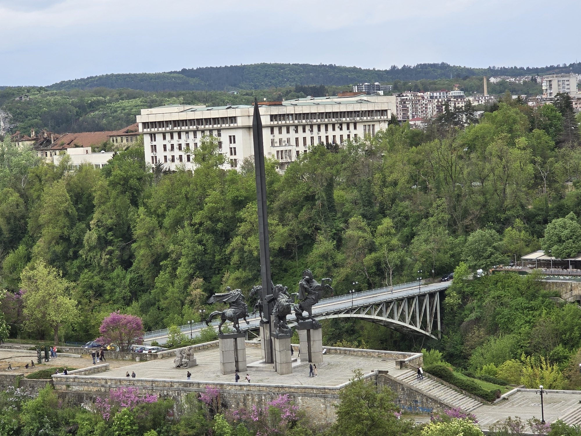The image shows a monument in Unknown, Unknown. The monument features four equestrian statues grouped around a tall, vertical sword. People are walking around the base of the monument. A bridge is visible in the background, behind the monument. Beyond the bridge, a large, light-colored building is situated on a wooded hill. The sky is overcast.