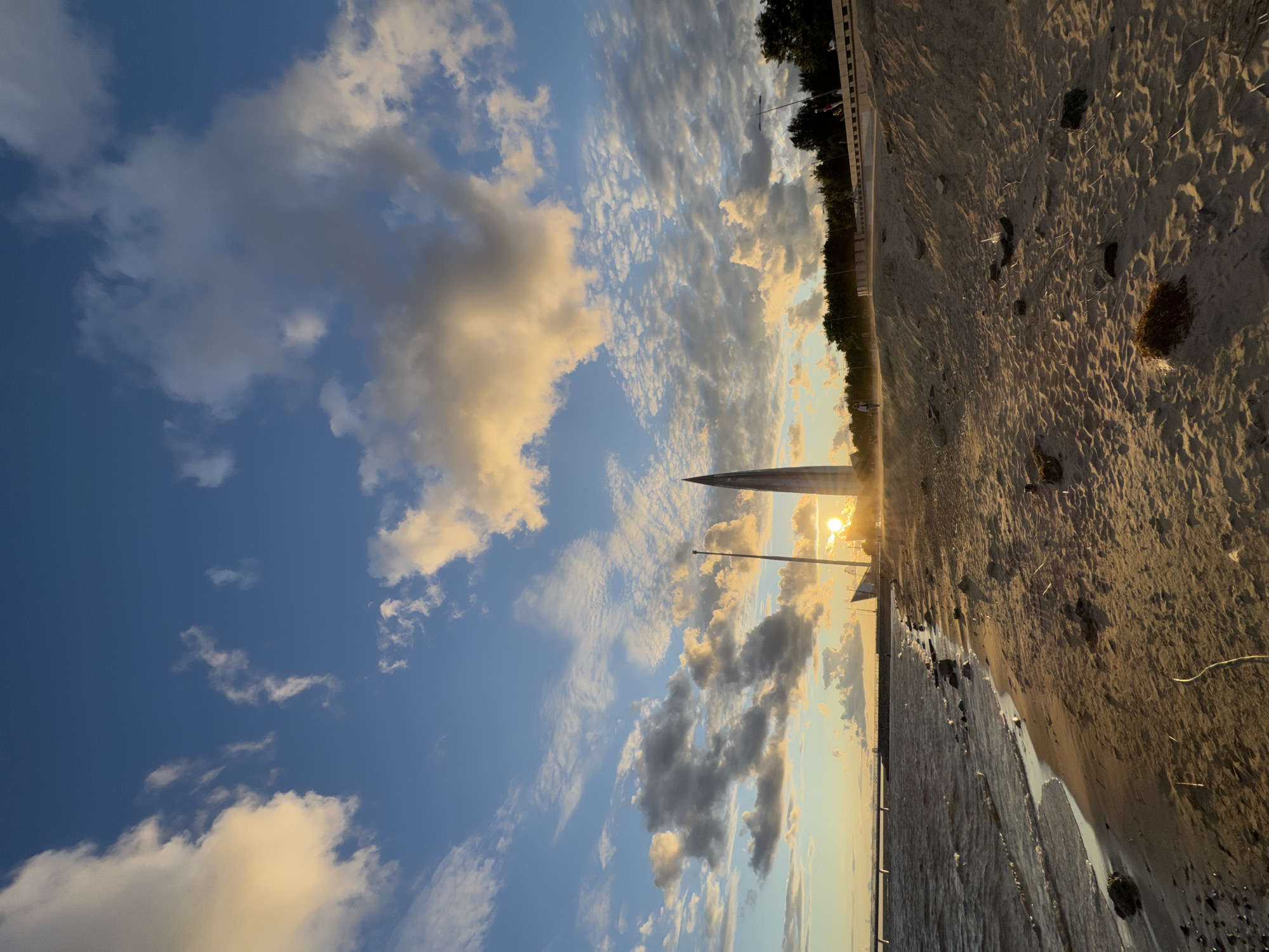 This is a vertical, eye-level, full shot of a beach next to a body of water during sunset. The sky is a vibrant blue with scattered clouds, some of which are illuminated by the setting sun, casting a warm, golden hue. In the distance, on the right side of the image, a tall, modern, glass skyscraper with a pointed spire dominates the skyline. To the left of the skyscraper, a tall, thin flagpole is visible. The sun is setting behind the skyscraper and flagpole, creating a strong glare. The beach is sandy with some scattered rocks and debris. The water is choppy with small waves lapping at the shore. A few people are visible on a walkway in the distance, and one person is walking on the beach. In the far distance, a long, low structure, possibly a pier or bridge, extends across the water. The overall impression is a serene and picturesque sunset scene in a coastal urban environment. This is the Lakhta Center in St. Petersburg, Russia.