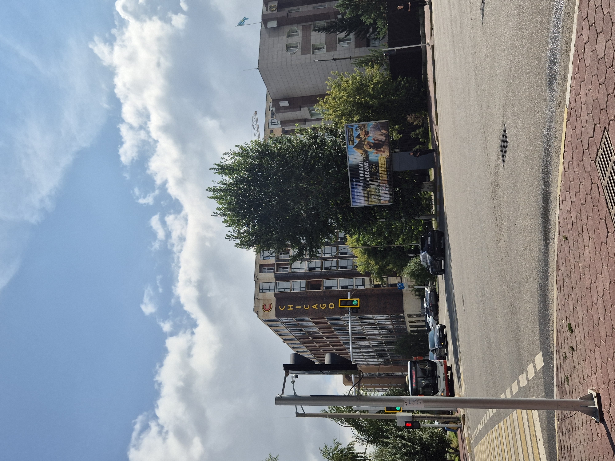 The image depicts a street scene in the city of Kokshetau, Kazakhstan. A multi-story building with the word "CHICAGO" visible on its facade is prominent in the background. The building appears to be a hotel or office complex, characterized by its brown brick and glass exterior. Adjacent to it, another building with a lighter facade, featuring distinctive arched windows, is visible. A billboard displaying an advertisement with images of people on a beach is situated between the two buildings. A street intersection is in the foreground, with traffic lights, a pedestrian crossing, and several vehicles, including a bus and cars. Trees line the street, and the sky is partially cloudy.