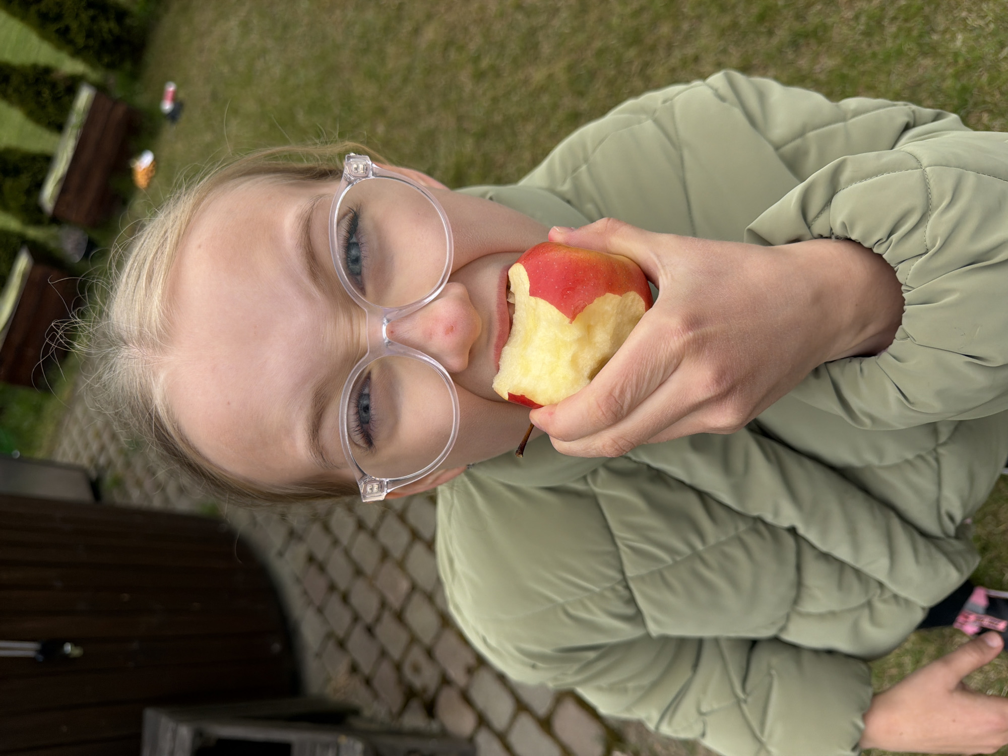 A young, fair-skinned girl with light blonde hair and blue eyes wearing clear-framed eyeglasses is shown from a close, elevated perspective. The girl is outdoors, appearing to be in a backyard with a lawn, indicated by the green grass and shrubs in the background. She is wearing a light green, padded jacket. The girl is holding a partially eaten red and yellow apple in her right hand and is in the process of eating it. The picture's orientation suggests it was taken with a phone held vertically. The location in Dreiliņi, Latvia, cannot be verified from the image.