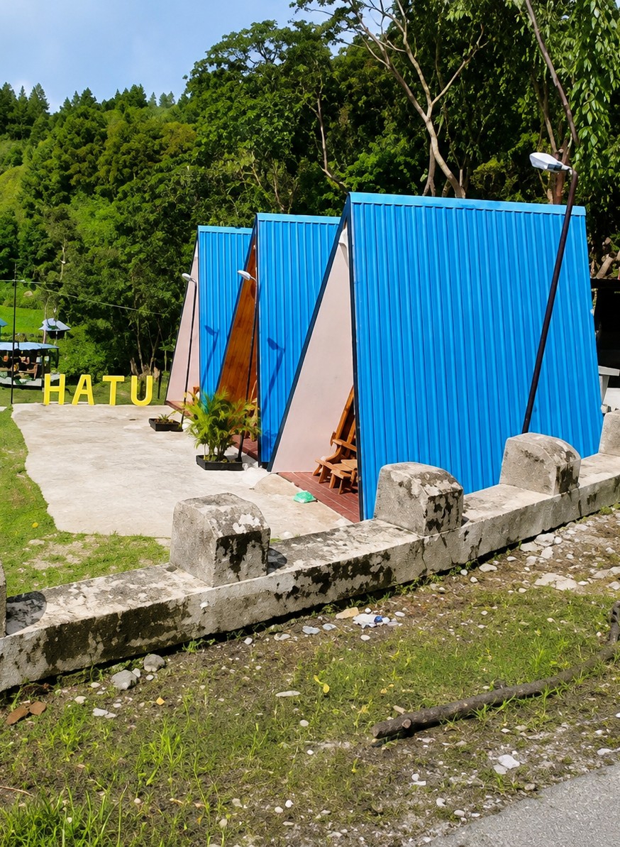 In Turpuk Sihotang, Indonesia, a row of three triangular, modern cabins with bright blue corrugated metal exteriors and light-colored triangular facades are positioned on a concrete ground. Yellow block letters spelling "HATU" are visible on the concrete in front of the cabins. Several potted plants, including a leafy green one, are situated near the entrance of the first cabin. Wooden benches or chairs are visible inside the openings of the cabins. A low, weathered concrete barrier with square posts runs across the foreground, separating the cabins from a grassy area with scattered rocks and debris, including plastic bottles and a tree branch. In the background, a lush, green forested hillside under a clear blue sky serves as the backdrop, with some bare tree branches extending over the cabins. The environment appears to be a resort or a similar recreational facility.