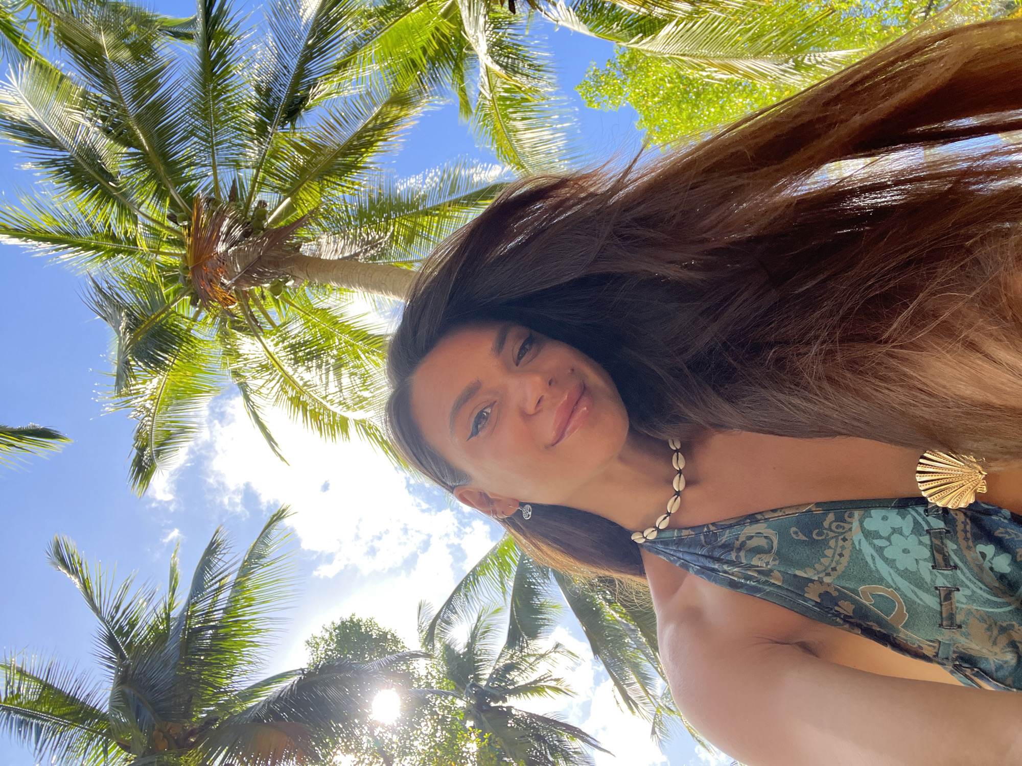 A woman with long, dark brown hair, wearing a teal patterned halter top with a gold seashell embellishment and a seashell necklace, smiles at the camera. She is outdoors, looking up at a bright blue sky filled with white clouds, framed by lush green palm trees. The scene appears to be a tropical location, possibly in the city of Koh Phangan, Thailand. The sun is shining brightly, casting a warm glow on her face and hair.
