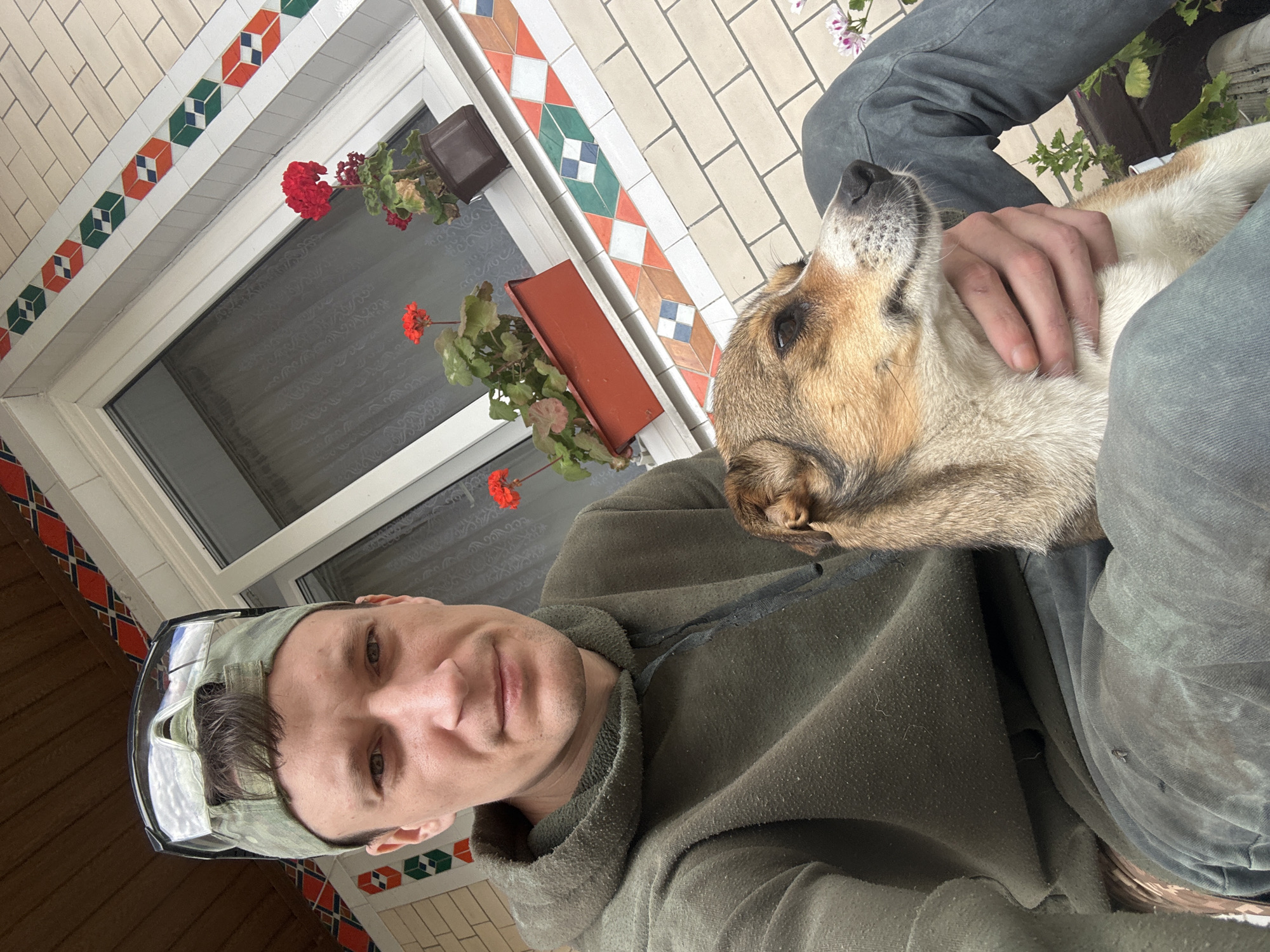 In a ground-level, outdoor, selfie-style photograph, a fair-skinned man with short brown hair is seated, holding a small-to-medium sized mixed-breed dog. The man is wearing a dark green hooded sweatshirt and dark pants. He has safety glasses perched on top of a camouflage baseball cap. The dog, with brown and white fur, is looking to the right, with the man's left hand supporting its chin and neck. They are positioned in front of a building with light-colored brickwork. Above the brick, a tiled decorative border runs along the base of a white-framed window. Two red potted geraniums are visible on the windowsill. The background suggests a residential area, consistent with the city of Makiv, Ukraine.