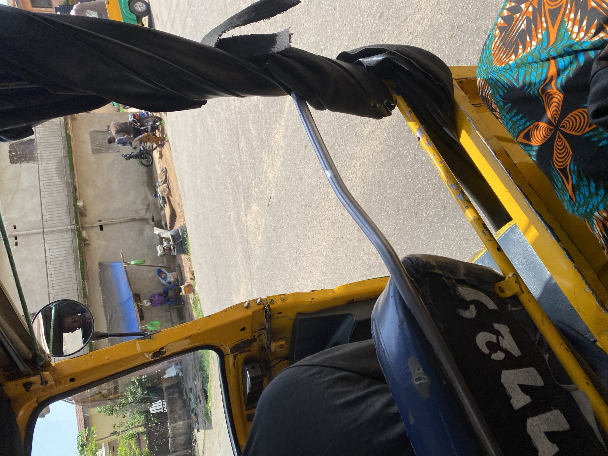 A man is seen from behind, wearing a black shirt and sitting in the driver's seat of a yellow auto-rickshaw. The auto-rickshaw has a dark blue seat with white numbers "7729" stenciled on it. The auto-rickshaw is on a paved road, with buildings and some vegetation visible in the background. In the distance, a man on a motorcycle is interacting with another man standing next to him. The scene appears to be in a residential or commercial area. The image is taken from inside the auto-rickshaw, looking forward and slightly to the right. The location is identified as Jos, Nigeria.