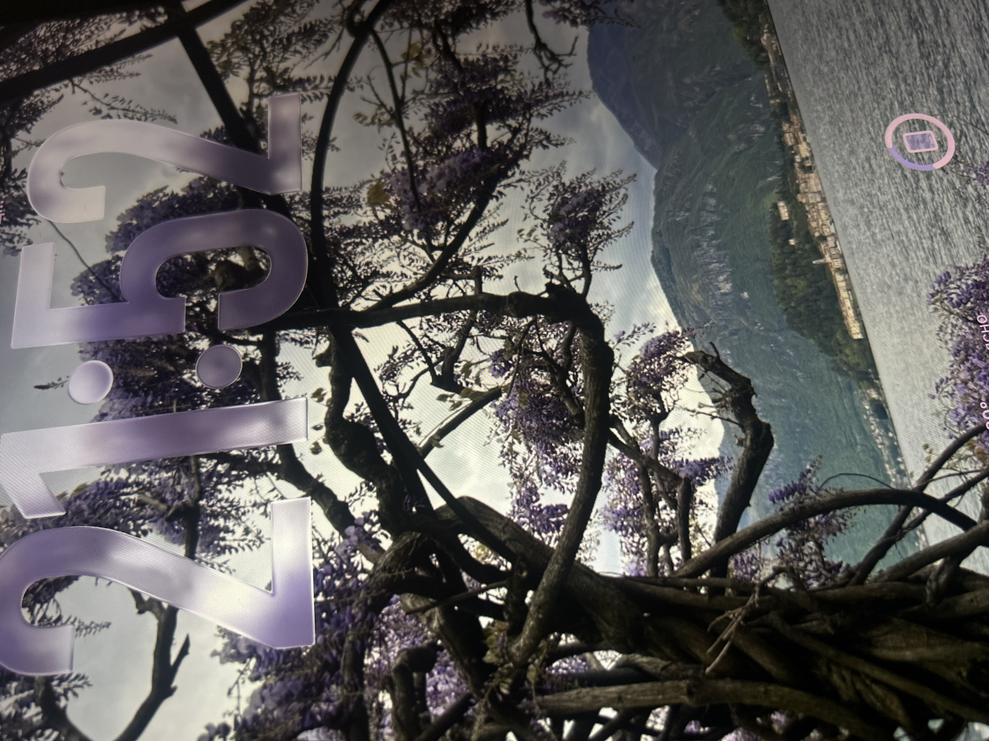 The image displays a close-up view of a digital clock showing the time "21:52", overlaid on a scenic background. The background features a large, gnarled tree with drooping purple wisteria flowers in the foreground. Behind the tree, a body of water is visible, with a picturesque town nestled along the shore and rolling green mountains in the distance under a cloudy sky. There is a circular icon with a square inside it in the lower right corner of the image. The image does not show the city of Krakow, Poland.
