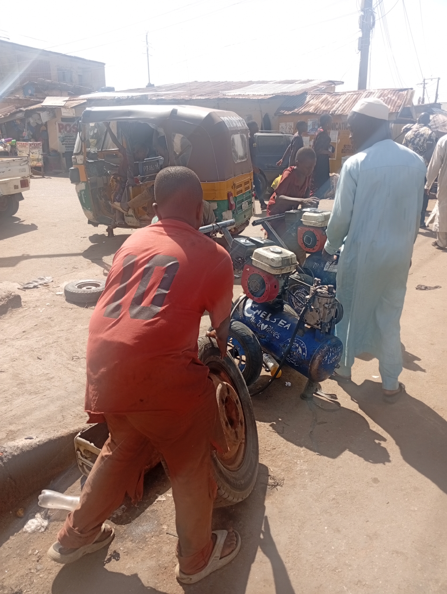The image depicts a street scene in Jos, Nigeria, with a man wearing an orange shirt with the number "10" on the back bending over a tire near the curb. A second man, wearing a light blue robe and a white cap, stands near a blue air compressor mounted on wheels. The air compressor has two red engines on top and the name "CHELSEA" written on the side. An auto-rickshaw is parked in the background, and several other people are walking in the vicinity. The environment suggests a busy street or market area with various buildings and structures visible in the background.